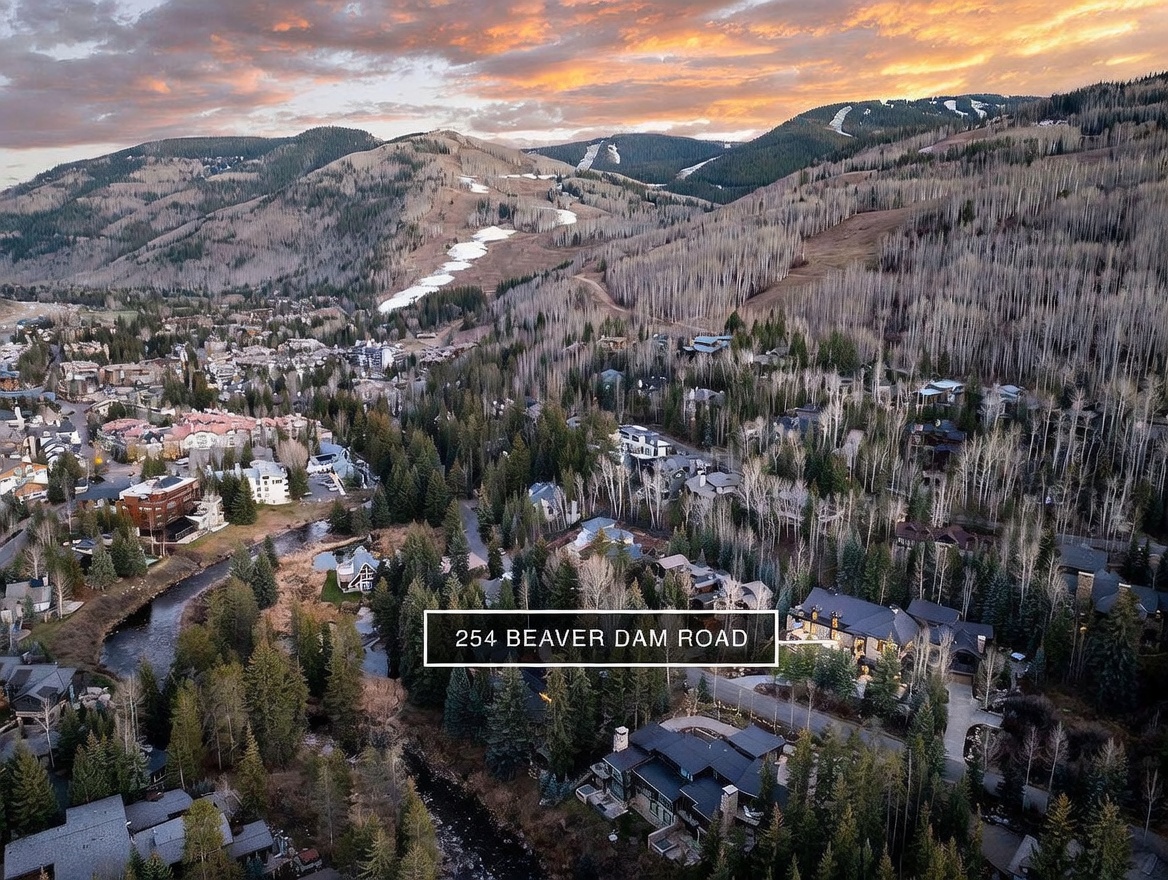 This high-angle aerial view captures a luxury mountain residence nestled within a dense forest of aspen and pine trees. The property is highlighted by a white circle, showcasing its proximity to a winding stream and a scenic ski resort backdrop under a dramatic sunset sky. The perspective emphasizes the home's secluded, natural setting and its integration into the mountainous landscape.
