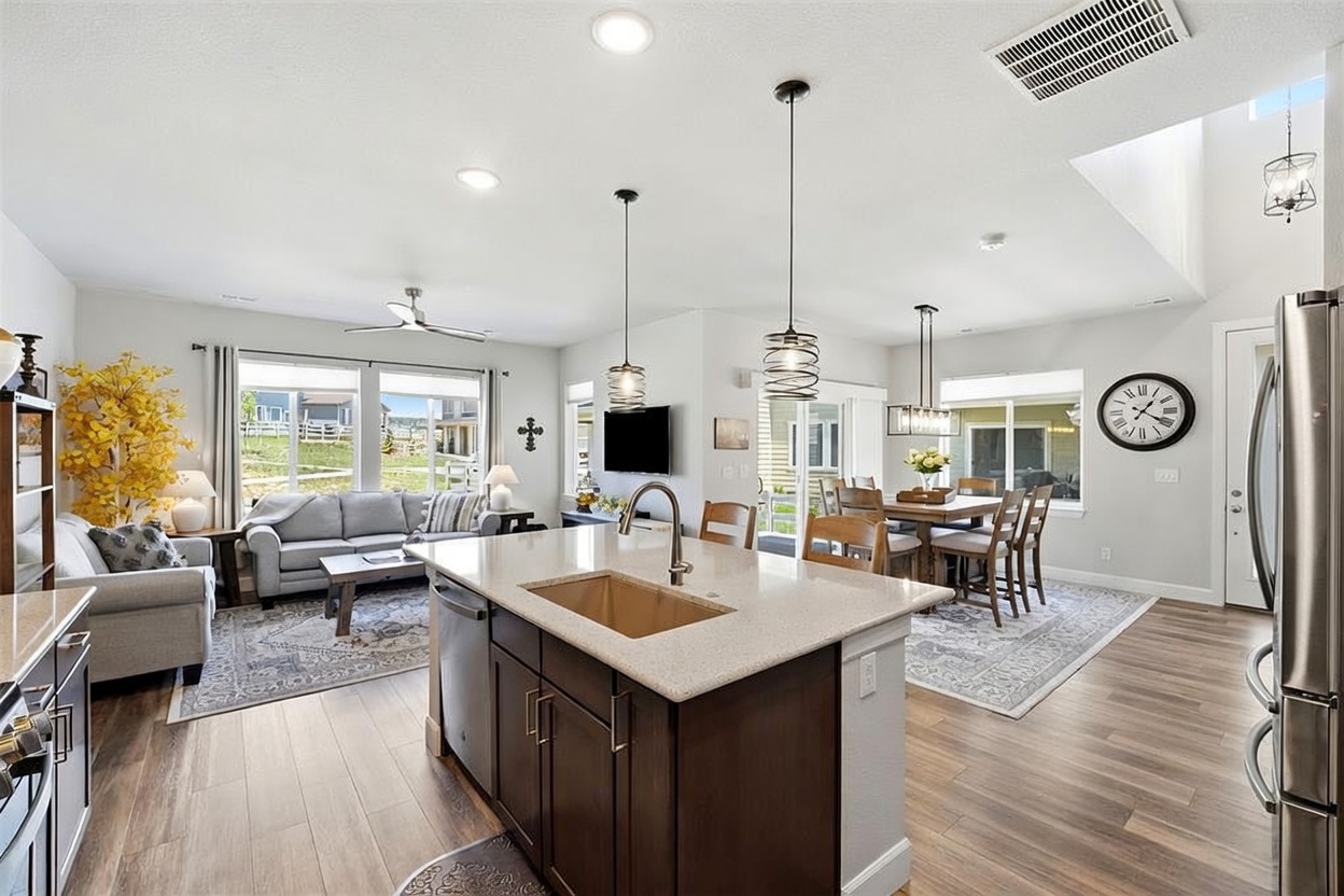 This open-concept living space features a central kitchen island with a sink and dark cabinetry, seamlessly flowing into a dining area and a comfortable living room. The interior is characterized by light-toned wood-style flooring, neutral wall colors, and a bright, airy atmosphere enhanced by natural light from large windows. The space is well-appointed with modern pendant lighting, stainless steel appliances, and a cohesive design that emphasizes functionality and a welcoming, contemporary aesthetic.
