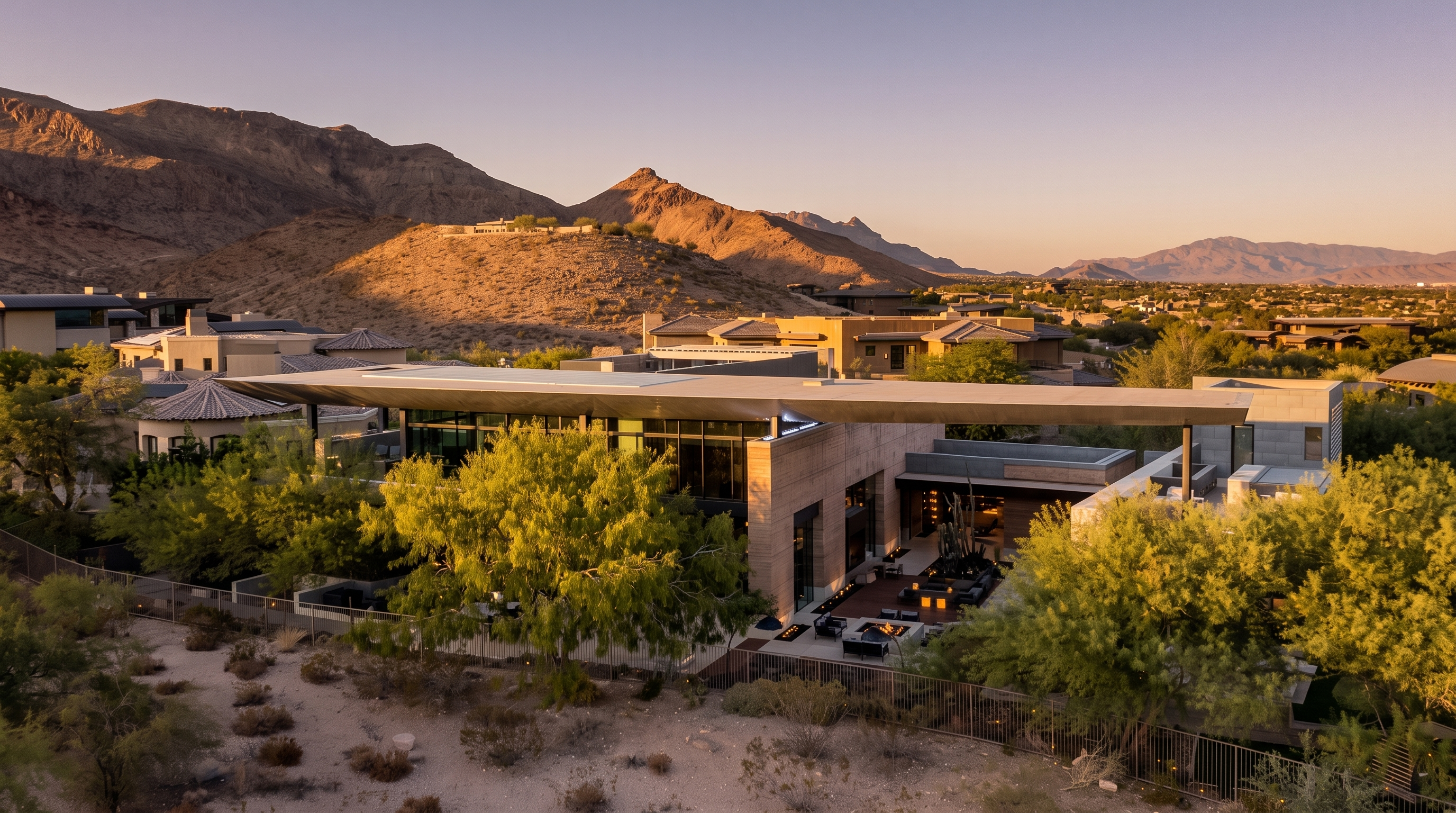 This aerial perspective captures a sprawling, modern luxury residence nestled against a dramatic desert mountain backdrop. The home features a prominent, flat, overhanging roofline that shades extensive glass walls and a sophisticated outdoor living area complete with a fire pit and lounge seating. The desert landscape, characterized by native vegetation and rocky terrain, seamlessly integrates with the architectural design, creating a serene and high-end atmosphere.