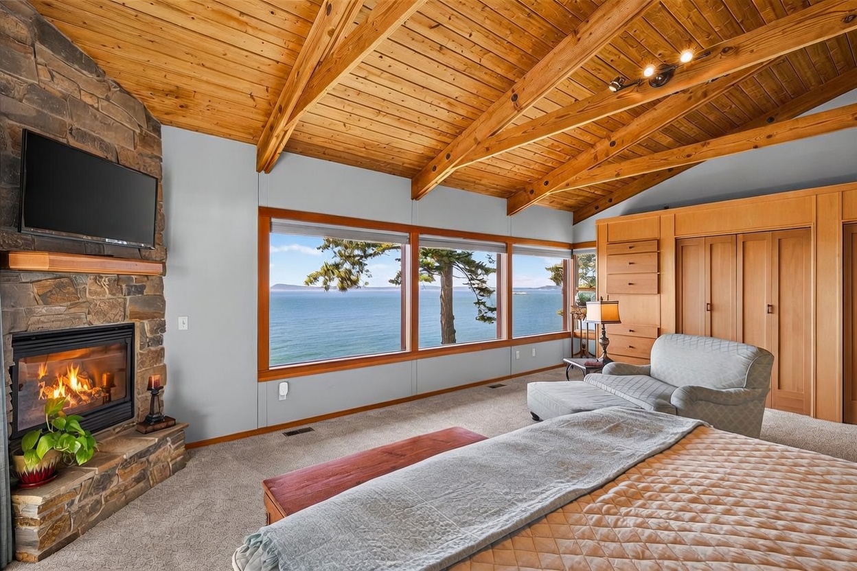 This primary bedroom features a vaulted wood-paneled ceiling with exposed beams and a stone-clad chimney breast housing a fireplace. A large expanse of windows provides expansive, unobstructed views of the ocean and distant coastline. The room is furnished with a comfortable seating area and built-in light wood cabinetry, creating a warm and serene atmosphere.