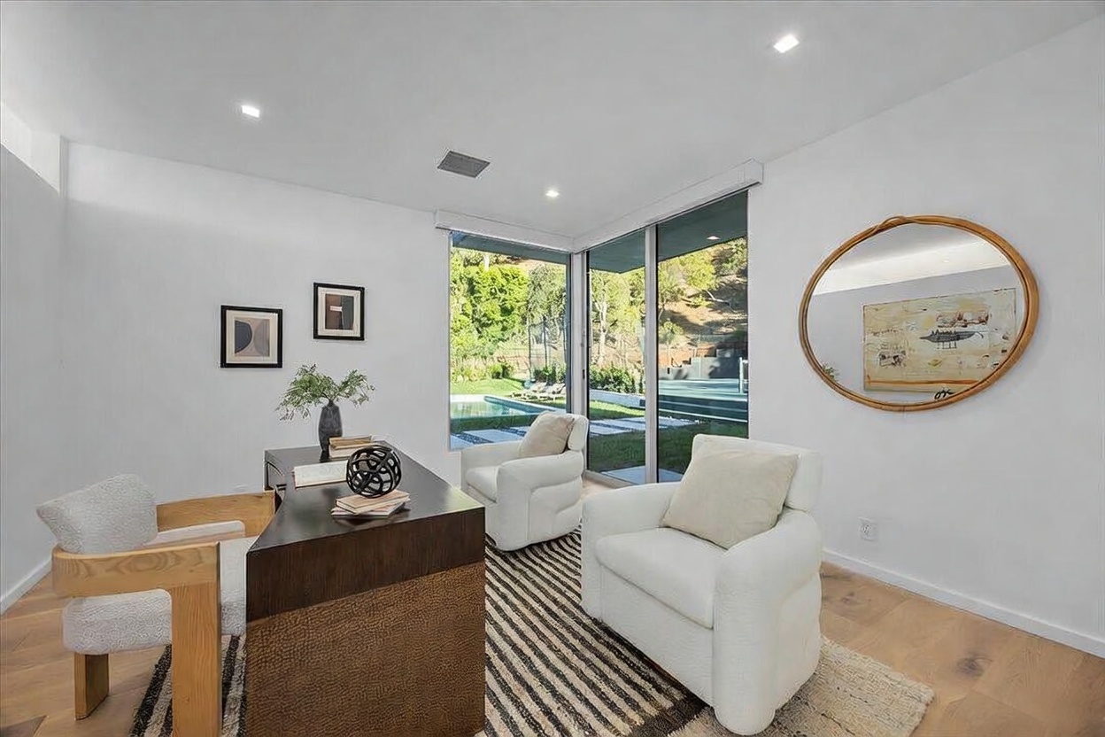 This modern home office features a sleek, dark-toned desk paired with a light wood chair and two comfortable white armchairs arranged on a striped area rug. Large glass sliding doors provide a seamless view of the outdoor pool and landscaped yard, filling the room with natural light. The space is accented by a large circular mirror and minimalist wall art, creating a sophisticated and professional atmosphere.