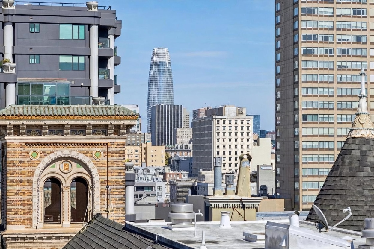 This elevated perspective captures a dense urban cityscape, highlighting a mix of historic and modern architectural styles. In the foreground, a detailed brick building with an arched window and decorative trim contrasts with the sleek, contemporary glass skyscraper rising prominently in the center. The scene provides a clear view of the surrounding skyline, emphasizing the property's central location within a bustling metropolitan area.