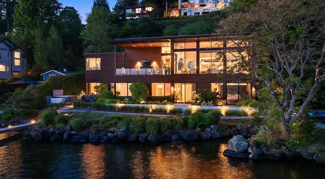 This stunning contemporary waterfront residence is captured at dusk, showcasing its modern architectural design and expansive glass walls that illuminate the interior. The home features a multi-level layout with a spacious balcony on the upper level and a landscaped terrace on the lower level, complete with a hot tub and integrated outdoor lighting. The perspective is taken from the water, highlighting the seamless connection between the home's sophisticated design and its serene lakeside setting.