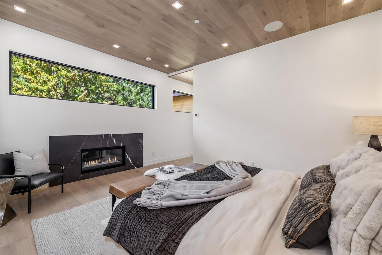 This modern primary bedroom features a striking black stone fireplace surround and a wide, horizontal window that frames views of the surrounding trees. The room is accented by a light wood-paneled ceiling with recessed lighting and a plush bed dressed in layered neutral textiles. A leather bench sits at the foot of the bed, contributing to the sophisticated and serene atmosphere of the space.