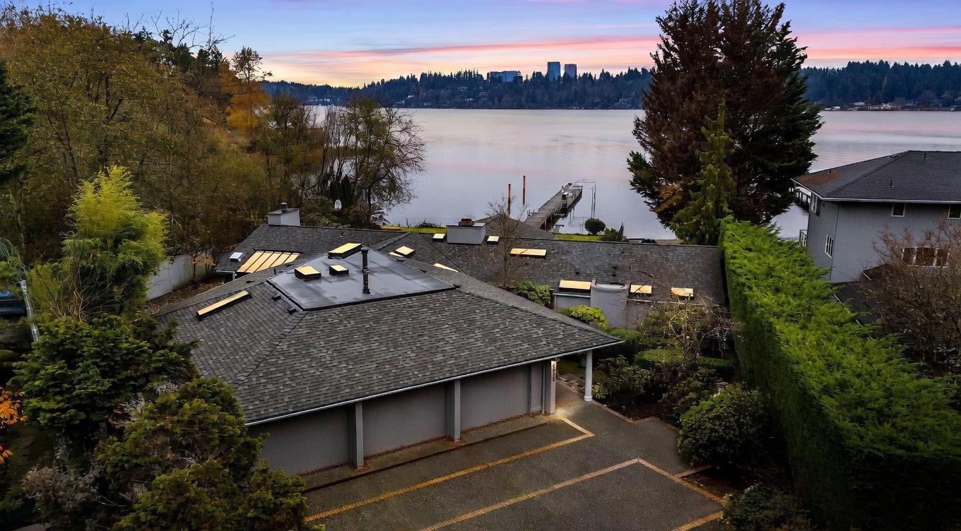 This elevated aerial perspective captures a waterfront property featuring a multi-faceted roofline with numerous skylights and a spacious paved driveway leading to a multi-car garage. The home is nestled among mature trees and lush landscaping, with a private dock extending into the calm lake beyond. The scene is set against a vibrant sunset sky, highlighting the serene and private nature of this lakeside estate.
