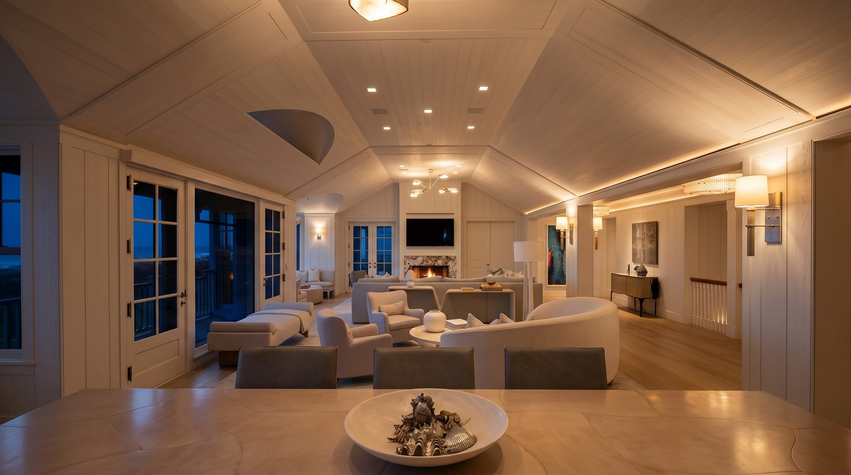 This expansive, light-filled living area features a vaulted ceiling with white paneling and a central pendant light, creating an airy and sophisticated atmosphere. The room is anchored by a comfortable seating arrangement, including a curved sofa and plush armchairs, centered around a stone-clad chimney breast with an integrated television. Large glass doors on the left provide a seamless transition to an outdoor space, while the foreground showcases a dining table topped with a decorative bowl of seashells.