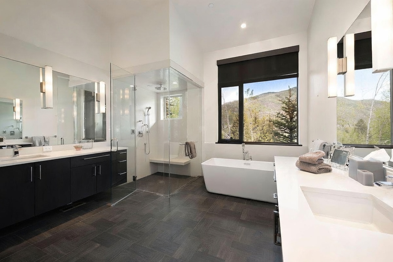 This spacious, modern primary bathroom features a double vanity with dark cabinetry, a large glass-enclosed walk-in shower, and a freestanding soaking tub positioned beneath a large window with mountain views. The room is finished with dark, textured floor tiles and crisp white walls, creating a clean and sophisticated aesthetic. The perspective captures the entire layout, highlighting the seamless integration of luxury fixtures and natural light.