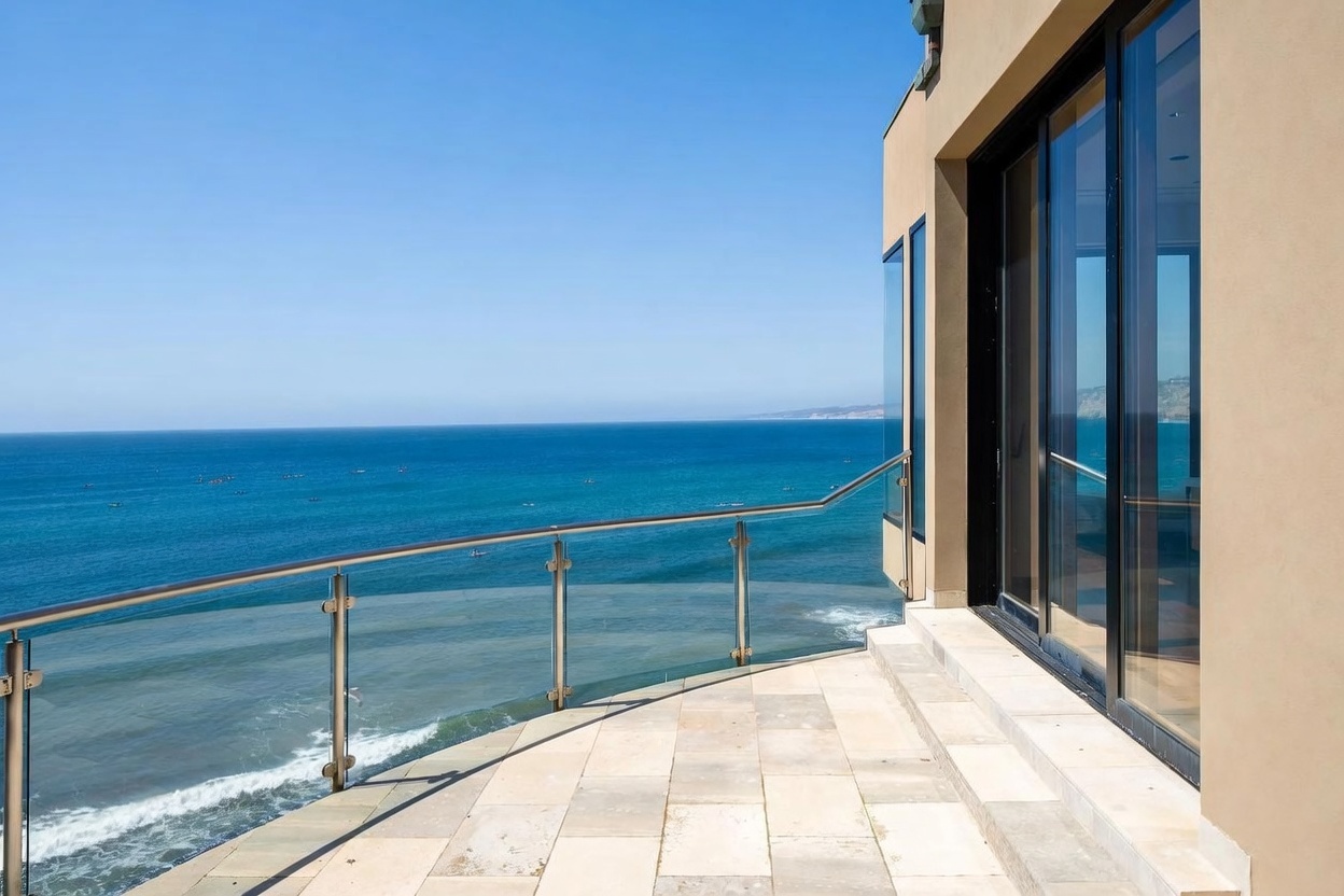 This balcony offers a stunning, unobstructed panoramic view of the ocean, featuring a sleek glass railing that maintains the visual connection to the horizon. The space is paved with light-toned stone tiles and provides direct access to the interior through large sliding glass doors. The perspective captures the seamless transition between the indoor living area and the expansive coastal scenery, emphasizing a luxurious, open-air lifestyle.