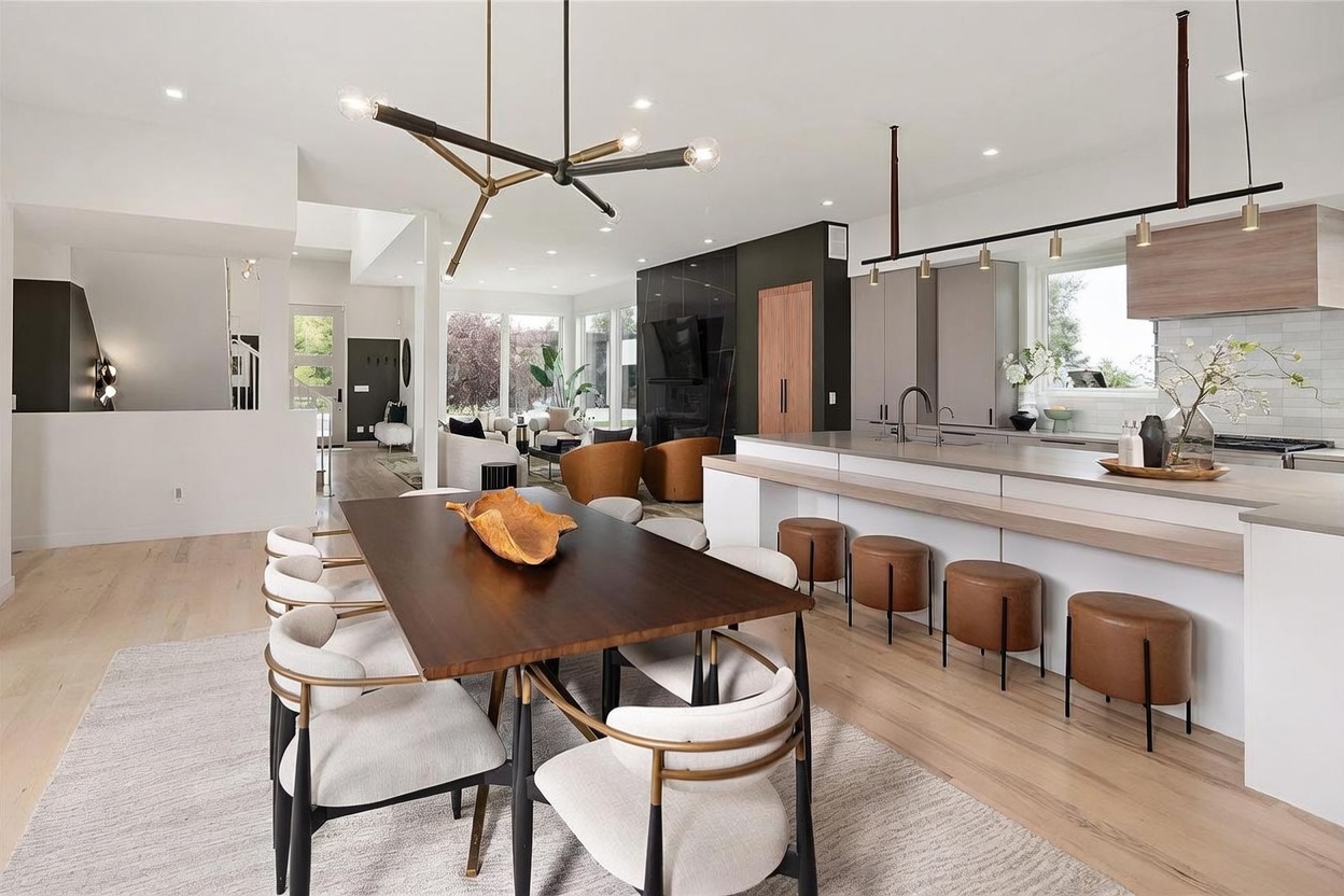 This open-concept living space features a modern kitchen with a large island, light wood cabinetry, and a sleek, minimalist aesthetic. A dining area with a dark wood table and upholstered chairs sits in the foreground, while a living area with warm-toned seating and a dark, tall fireplace surround is visible in the background. The room is illuminated by a striking geometric chandelier and recessed lighting, creating a bright and inviting atmosphere.