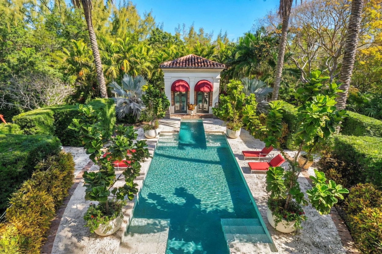 This high-angle shot captures a serene, resort-style swimming pool surrounded by lush tropical landscaping and manicured hedges. At the far end of the pool stands a classic pavilion with red fabric awnings over its glass-paned doors, creating a sophisticated focal point. The scene is completed by stone decking, vibrant potted plants, and red lounge chairs, evoking a luxurious and private outdoor retreat.