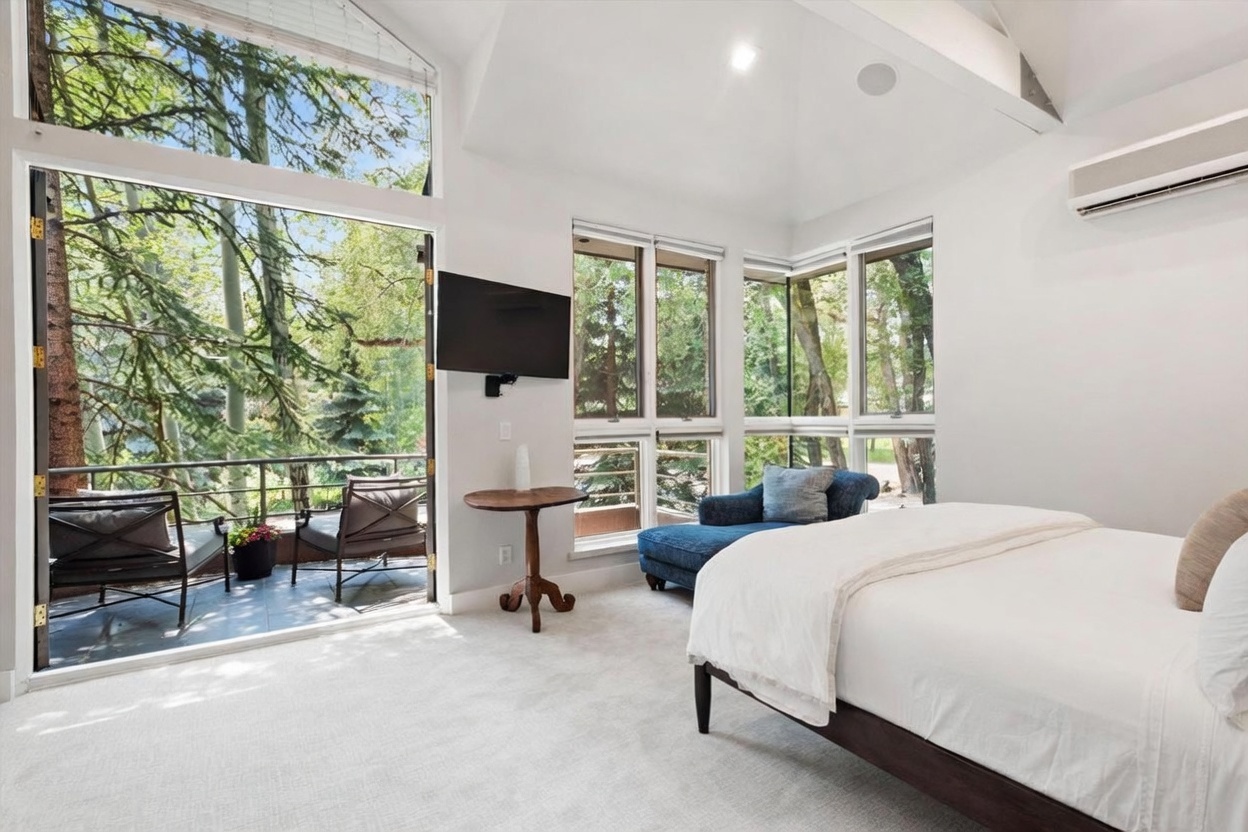 This bright and airy primary bedroom features a neutral color palette and expansive windows that offer a seamless connection to the outdoors. The room includes a comfortable bed, a cozy blue chaise lounge tucked into a corner, and a small wooden side table beneath a wall-mounted television. A glass door opens directly onto a private balcony, enhancing the sense of space and providing a serene view of the surrounding trees.