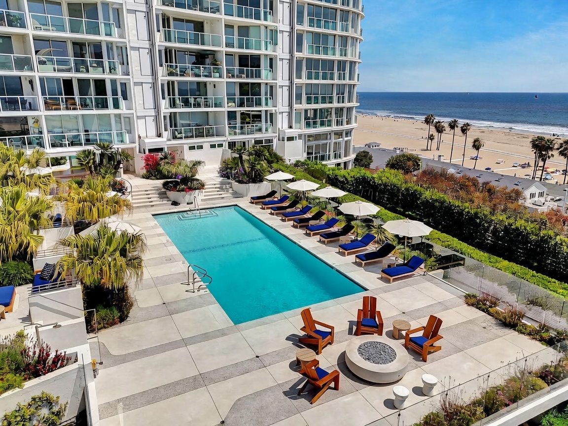 This high-angle aerial view captures a luxurious resort-style pool deck overlooking a sandy beach and the ocean. The space features a rectangular swimming pool lined with lounge chairs and umbrellas, complemented by a circular fire pit area with wooden seating. The modern multi-story building provides a sleek backdrop, emphasizing the property's prime coastal location and upscale outdoor living amenities.