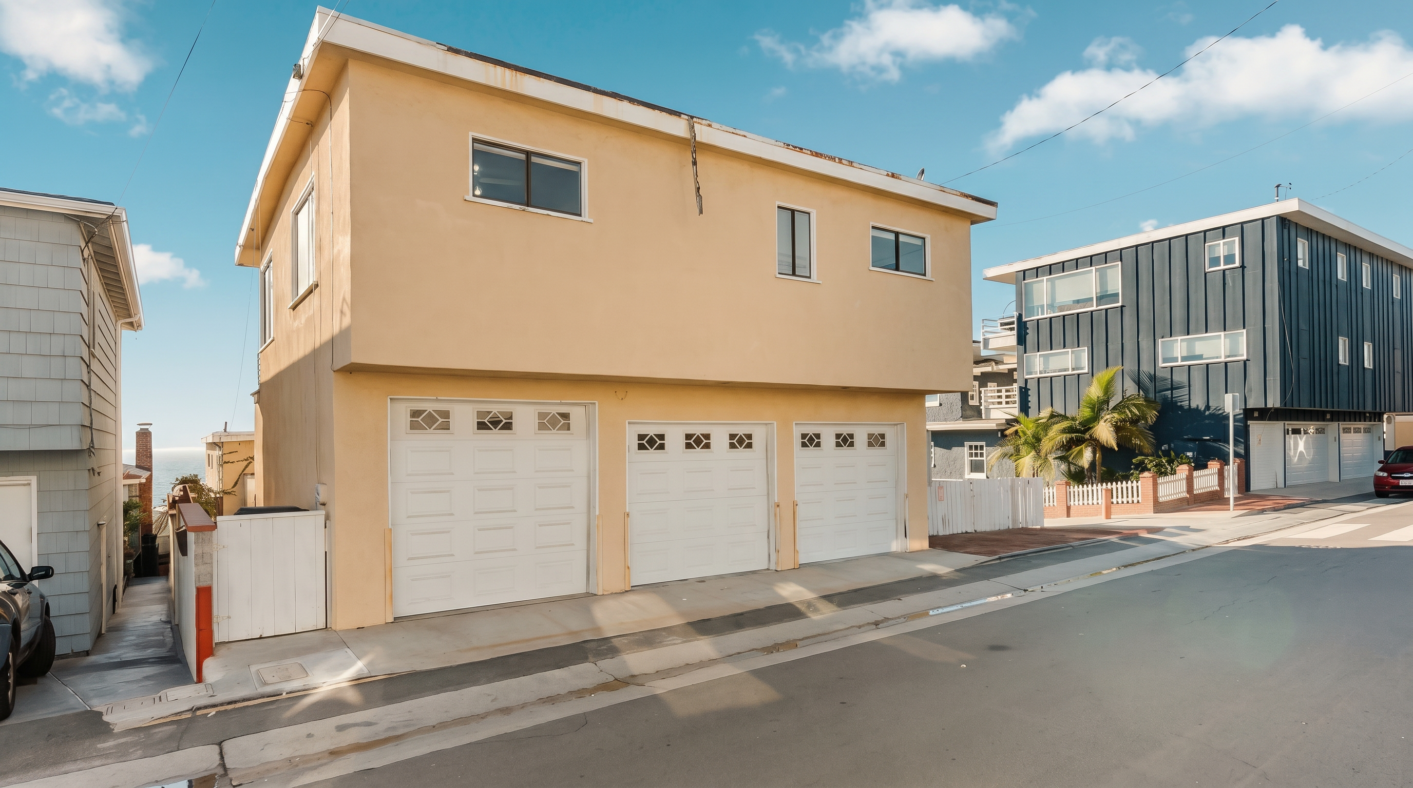 This exterior view captures a two-story residential building with a tan stucco facade, featuring three white garage doors on the ground level. The structure is positioned on a slight incline along a paved street, with a narrow walkway leading to the side of the property. The perspective is from the street level, showcasing the building's simple architectural lines and its proximity to neighboring homes under a bright, partly cloudy sky.