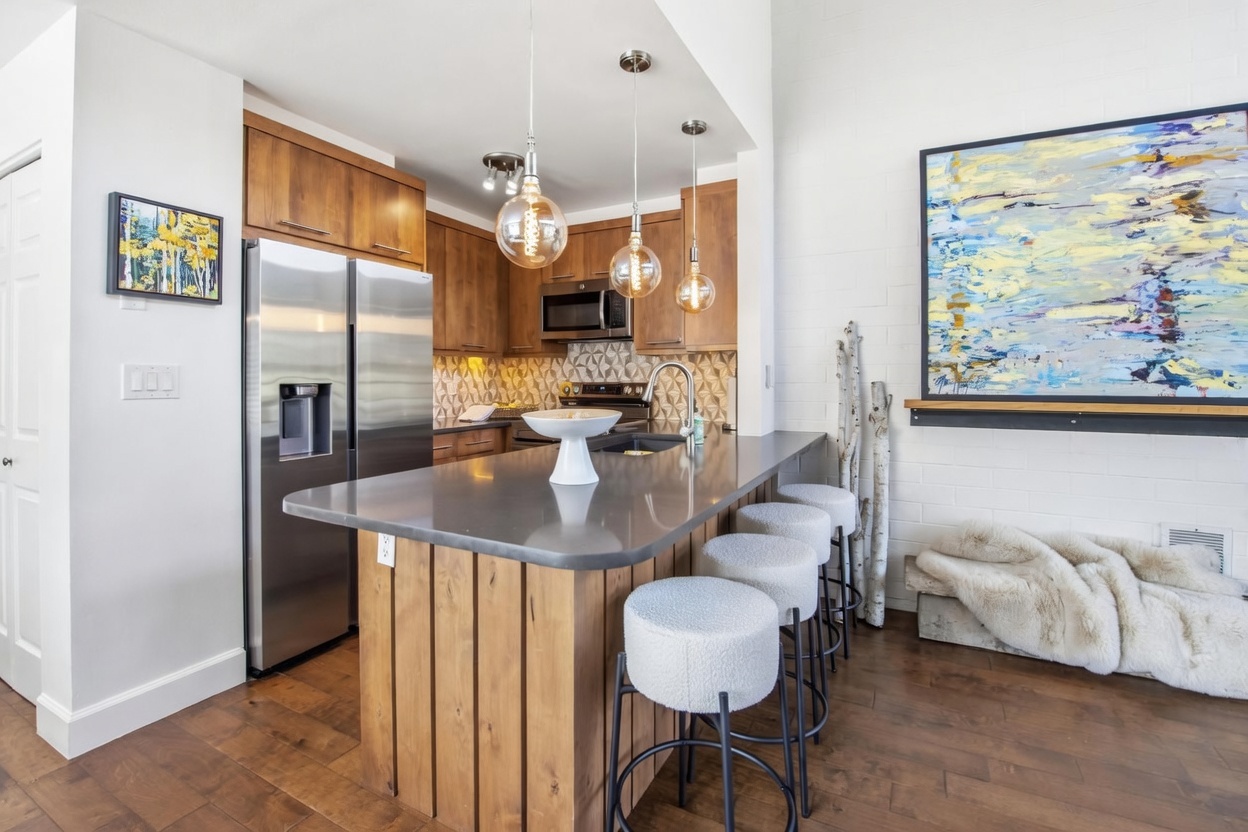 This modern kitchen features a central island with a dark countertop and vertical wood paneling, complemented by three textured bar stools. The space is equipped with stainless steel appliances, warm wood cabinetry, and a geometric patterned backsplash. A large, vibrant abstract painting hangs on the adjacent white brick wall, adding a bold artistic touch to the cozy, well-lit area.