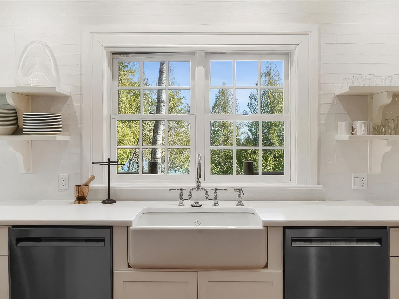 This bright, clean-lined kitchen features a classic farmhouse sink centered beneath a large, multi-pane window that offers a view of lush green trees. The space is framed by white subway tile backsplash and symmetrical open shelving on either side, with dark stainless steel appliances flanking the sink area. The overall aesthetic is airy and traditional, emphasizing a functional, light-filled workspace.