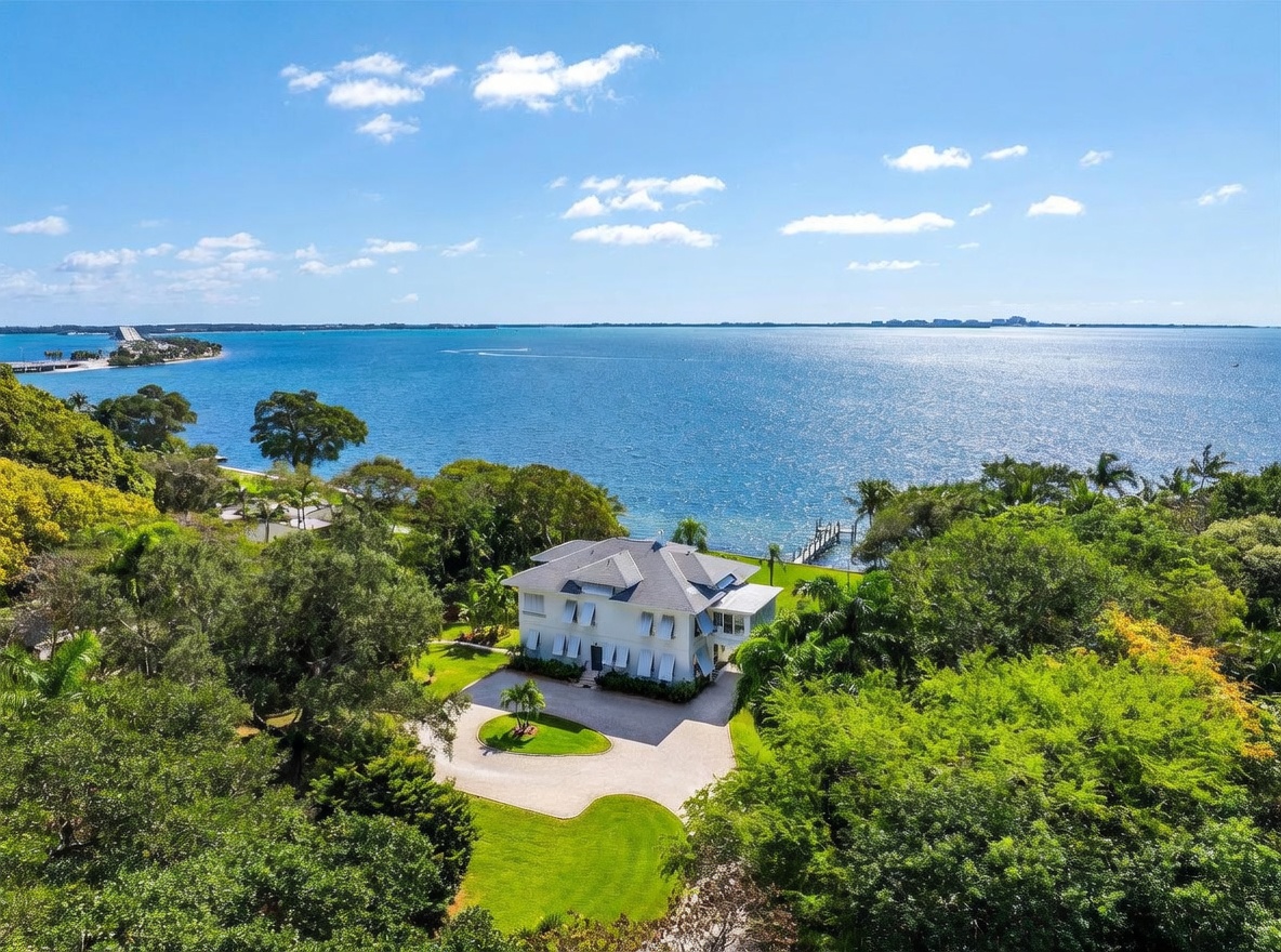 This high-angle aerial shot captures a stately, multi-story white residence situated on a lush, expansive waterfront property. The home features a classic architectural design with a circular driveway, surrounded by mature trees and manicured green lawns that lead directly to the water's edge and a private dock. The expansive blue bay stretches into the horizon, creating a serene and prestigious coastal atmosphere.