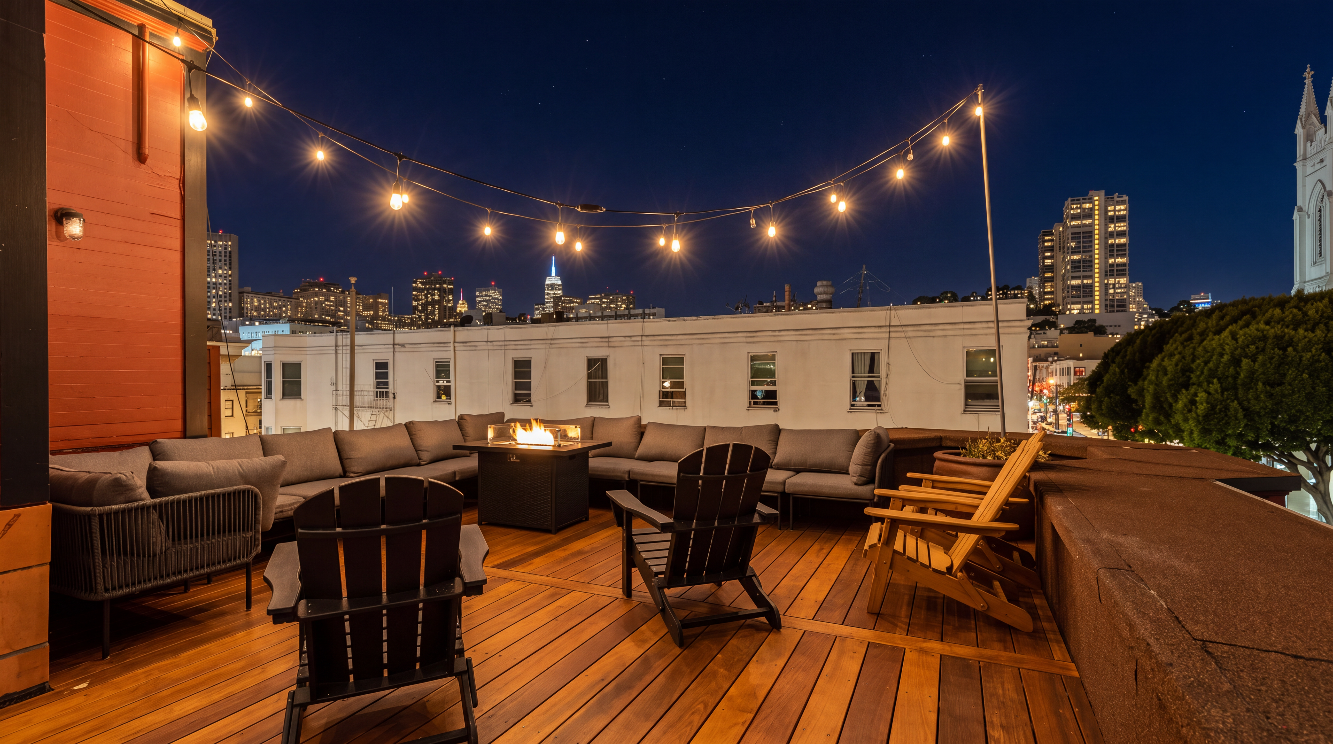 This stunning rooftop deck offers a sophisticated outdoor living space complete with a modern fire pit, plush modular seating, and dark wooden Adirondack chairs. String lights create an inviting ambiance, while the panoramic city skyline view provides a dramatic backdrop. It is an ideal setting for evening entertaining and urban relaxation.