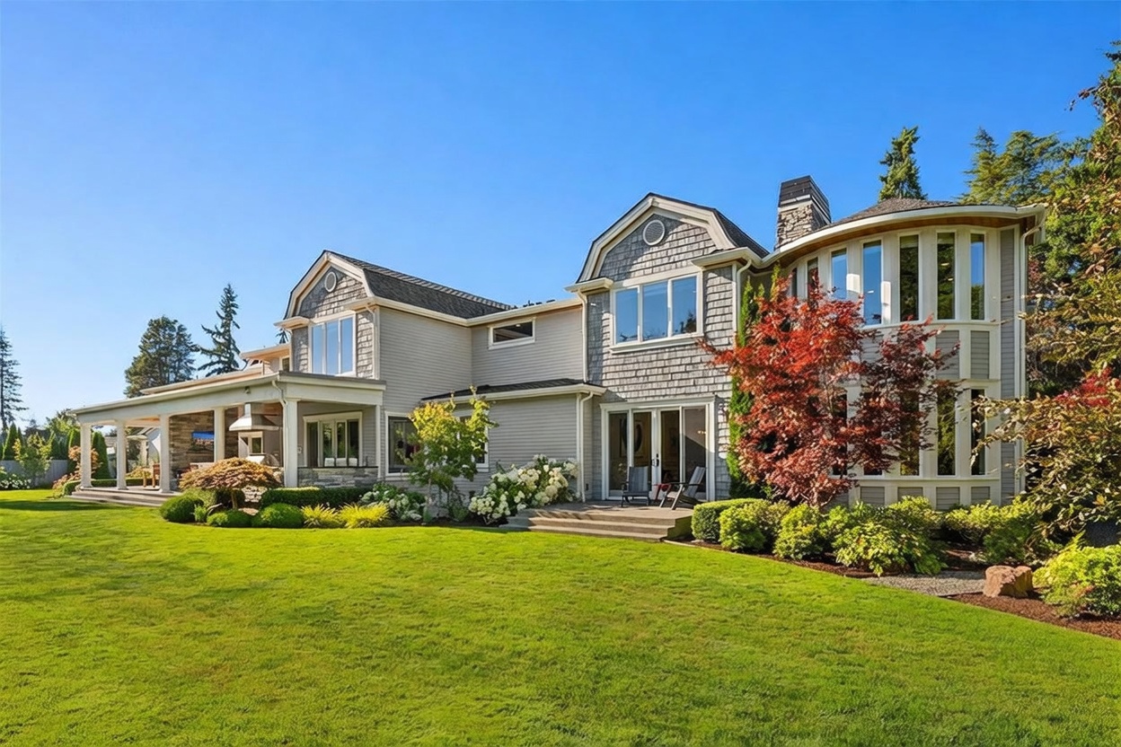 This rear view of a large, multi-story home showcases a blend of horizontal siding and shingle-style siding, featuring a prominent curved architectural section with tall windows. A spacious covered patio area with stone pillars provides an outdoor living space, while a well-manicured lawn and mature landscaping frame the property. The bright, clear blue sky highlights the home's detailed rooflines and expansive windows, creating an inviting and upscale residential atmosphere.