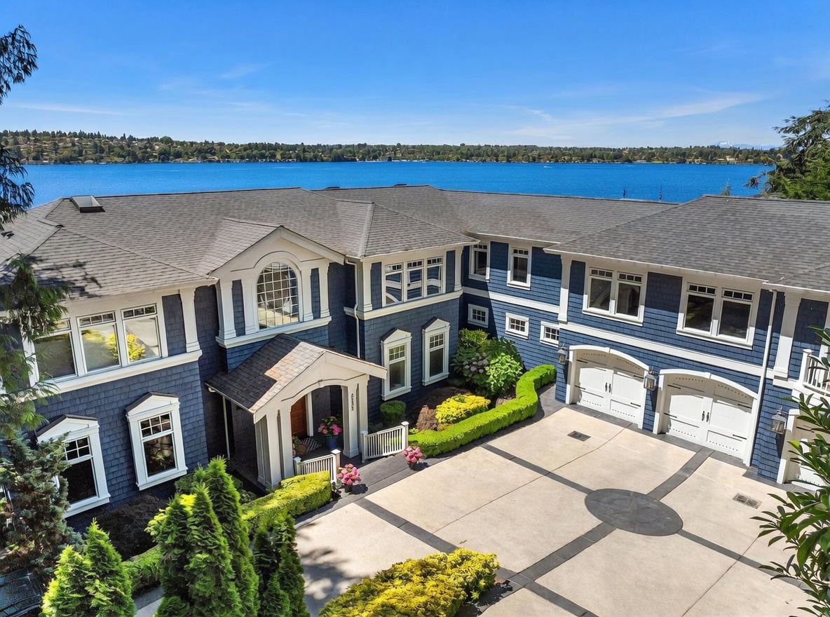 This high-angle aerial shot captures a stunning, large blue-shingled waterfront estate with a prominent front entryway and a multi-car garage. The property features a well-manicured garden with neatly trimmed hedges and a spacious concrete driveway, all set against the backdrop of a vast, sparkling blue lake under a clear sky. The architectural style is elegant and traditional, emphasizing luxury living with expansive views.