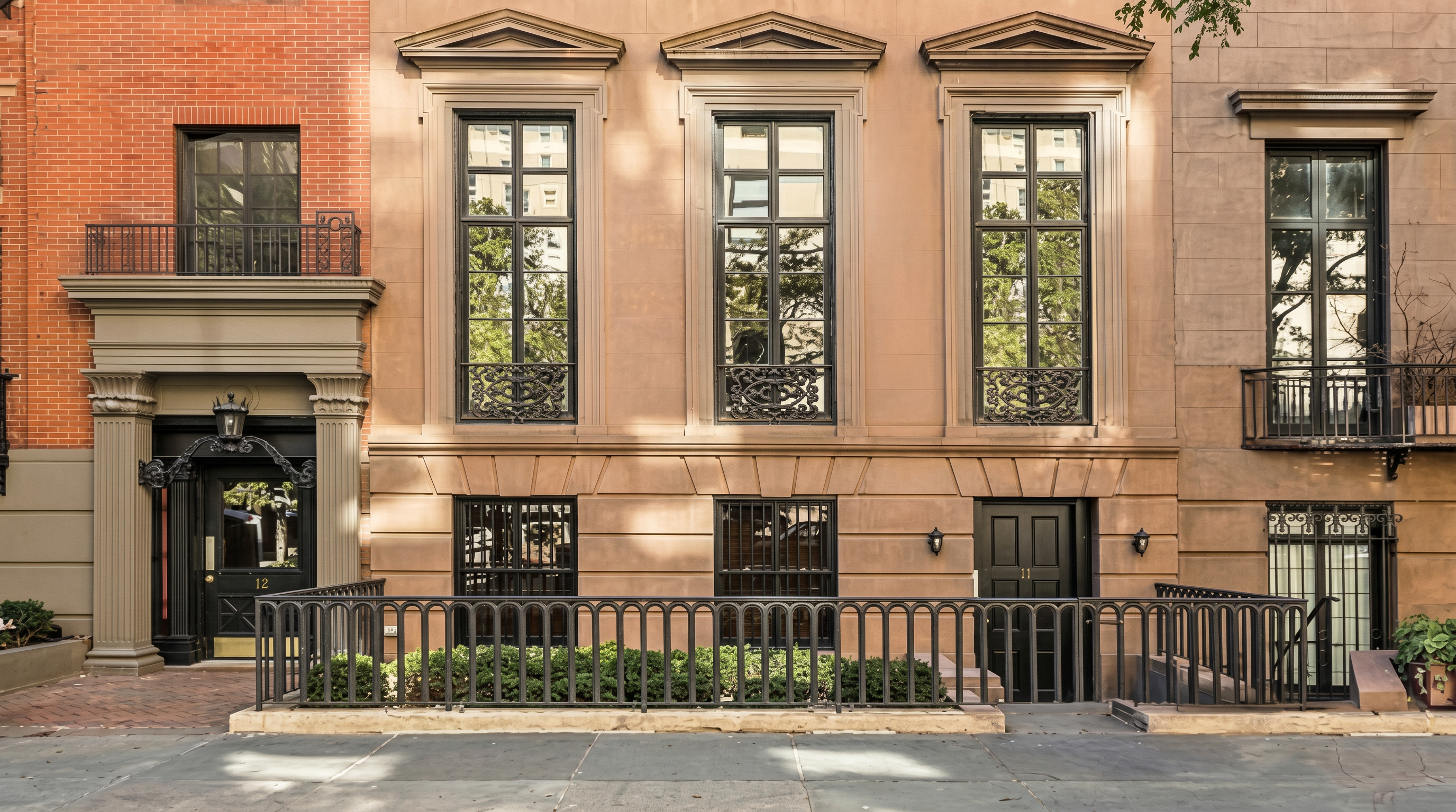 This stunning brownstone townhouse features a classic architectural design with large, symmetrical windows and ornate iron railings. The ground floor entrance is highlighted by an elegant doorway and a manicured hedge behind a traditional black metal fence. The overall appearance is sophisticated and quintessentially upscale, conveying a sense of historic charm and urban luxury.