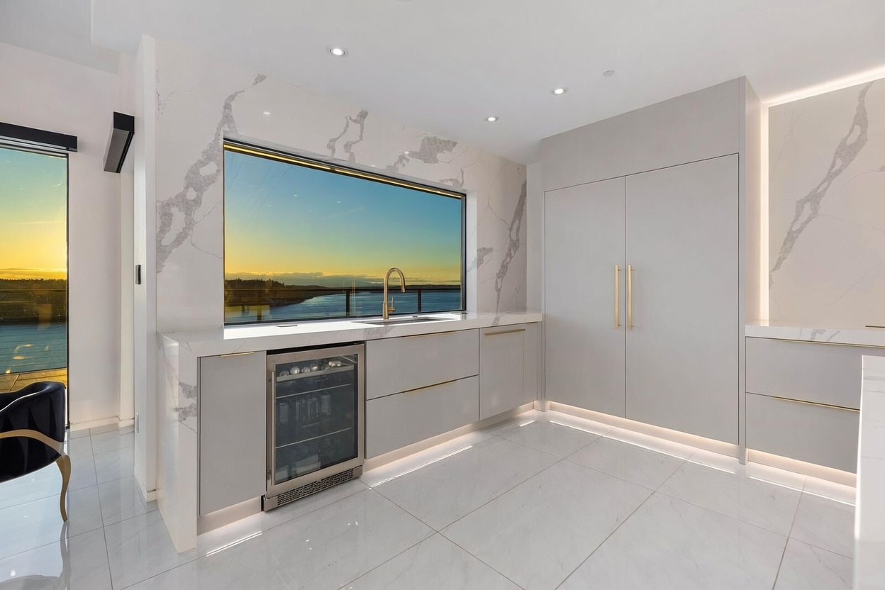 This sleek, modern kitchen features light-toned cabinetry with gold-tone hardware and a large, integrated wine refrigerator. A wide, rectangular window provides a stunning sunset view over the water, framed by a continuous stone backsplash that extends across the wall. The space is finished with polished, light-colored tile flooring and under-cabinet lighting, creating a bright and sophisticated atmosphere.