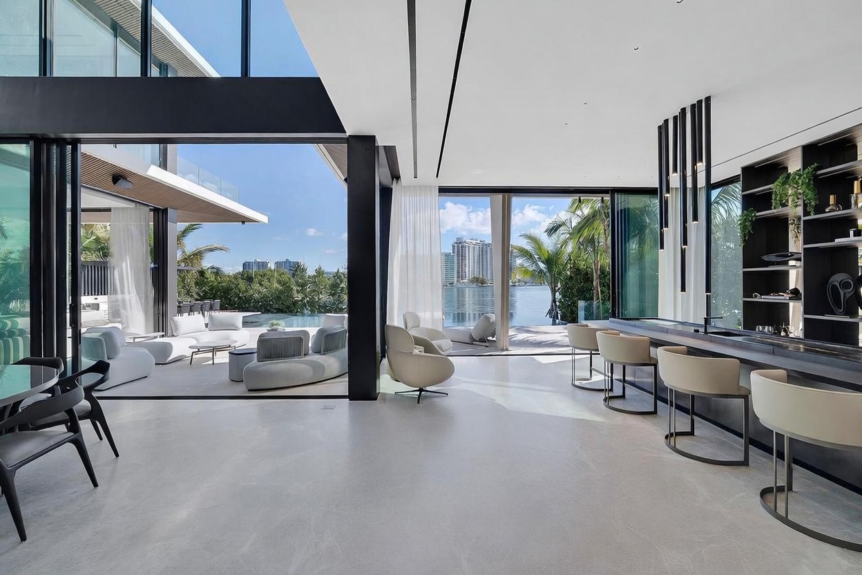 This expansive, modern living space features an open-concept design with seamless transitions to an outdoor patio and pool area through large glass sliding doors. The interior includes a sleek bar area with contemporary stools and minimalist pendant lighting, set against a backdrop of polished light-toned flooring. The room offers a panoramic view of the waterfront and city skyline, creating a sophisticated and airy atmosphere.