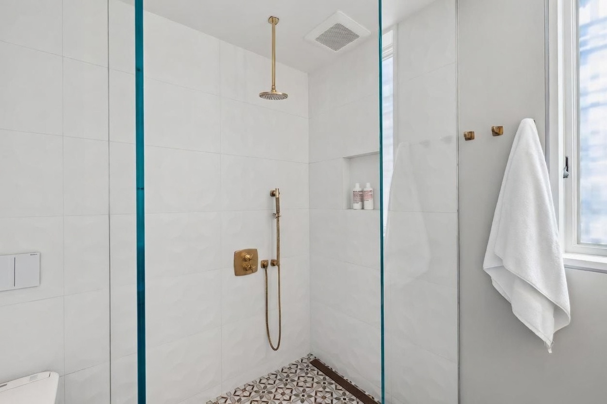 This modern walk-in shower features crisp white wall tiles and a striking patterned tile floor, accented by elegant gold-toned fixtures including a rainfall showerhead and handheld sprayer. A glass partition separates the shower from the rest of the bathroom, while a recessed niche provides convenient storage for toiletries. The space is bright and clean, with a white towel hanging on a nearby wall hook, creating a polished and contemporary aesthetic.