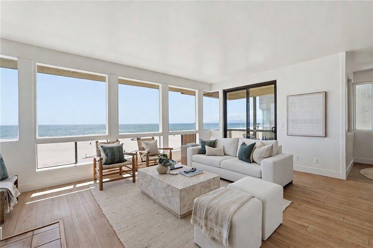 This bright and airy living room features a neutral color palette and expansive windows that provide a panoramic view of the ocean and beach. The space is furnished with a comfortable white sofa, two wooden armchairs with patterned cushions, and a large, light-toned stone coffee table centered on a textured area rug. The light wood flooring and minimalist decor create a serene, coastal-inspired atmosphere perfect for relaxing while overlooking the water.
