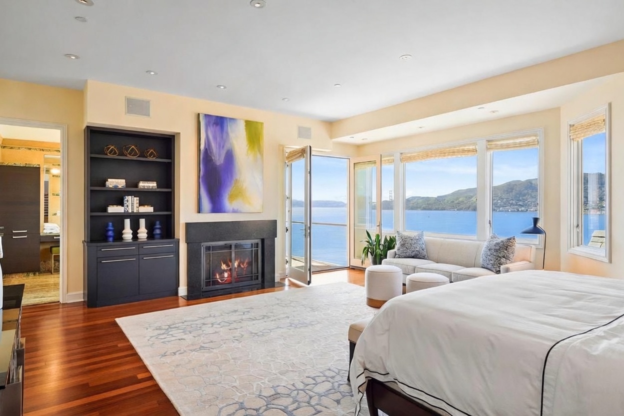 This spacious primary bedroom features rich hardwood flooring and a large area rug, centered around a dark-toned fireplace with a prominent abstract painting above. A comfortable seating area is positioned near expansive windows that provide panoramic water views and access to a private balcony. The room is bathed in natural light, creating a serene and luxurious atmosphere with a view of the distant hills.