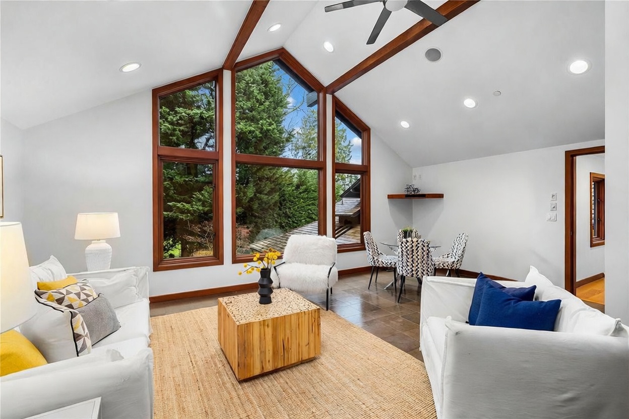 This bright and airy living room features a dramatic vaulted ceiling with exposed wooden beams and a large, multi-pane window wall that brings in abundant natural light. The space is furnished with comfortable white sofas, a unique wooden block coffee table, and a cozy dining area in the corner, creating an inviting open-concept layout. The combination of modern decor, warm wood accents, and expansive views of the surrounding greenery gives the room a sophisticated yet relaxed atmosphere.