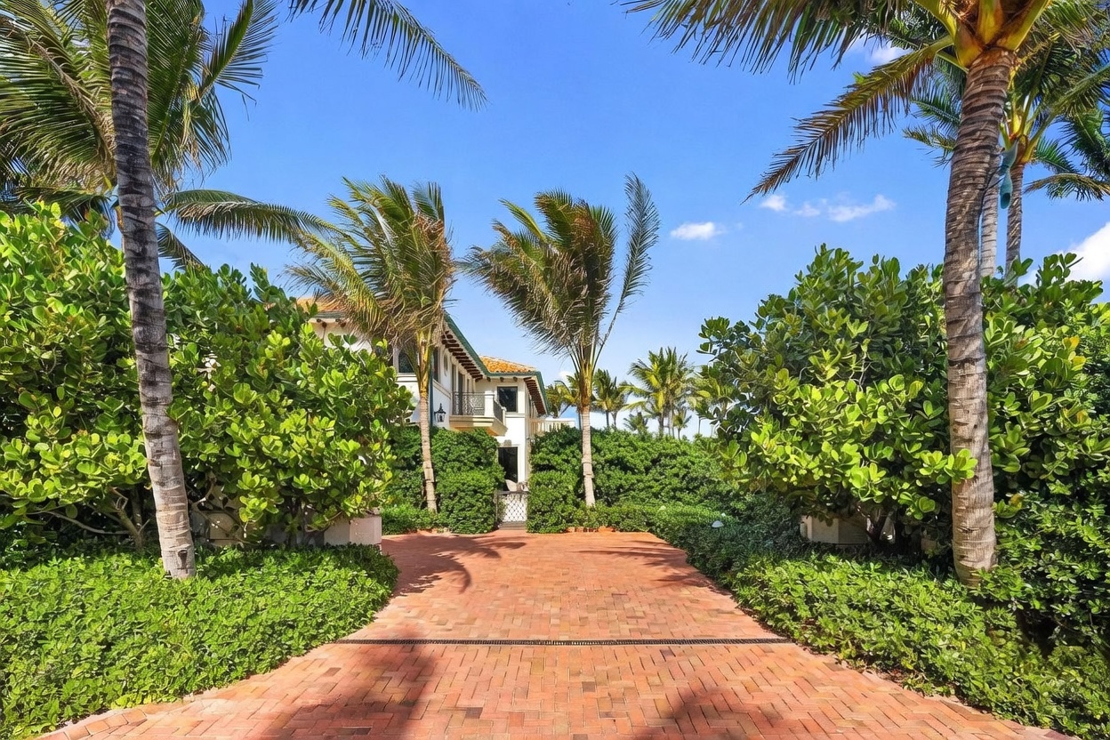 This image captures a brick-paved driveway leading toward the entrance of a multi-story residence surrounded by lush tropical landscaping. Tall palm trees frame the view, creating a sense of privacy and elegance as the path winds toward the home's white facade. The bright blue sky and vibrant greenery contribute to a serene, high-end coastal atmosphere.