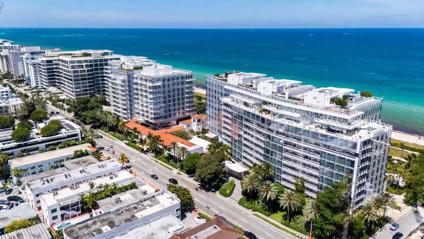 This high-angle aerial view captures a luxury coastal residential complex situated directly along a turquoise oceanfront. The scene features multiple modern, multi-story buildings with extensive glass facades and flat roofs, surrounded by lush tropical landscaping and palm trees. A coastal road runs between the buildings, providing a clear perspective of the property's prime location and proximity to the beach.