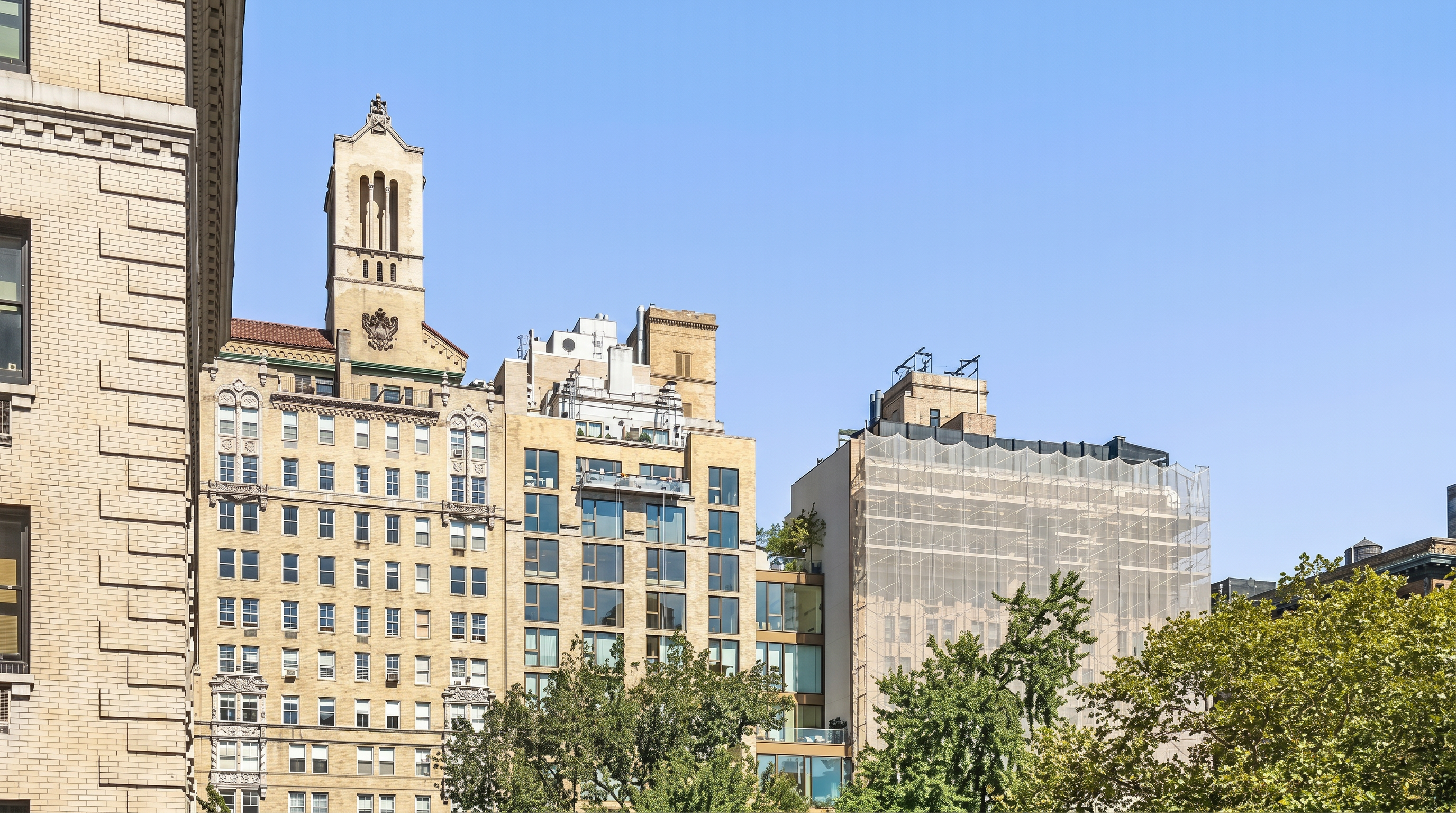 This street-level view shows a collection of classic and modern urban apartment buildings set against a clear blue sky. The prominent centerpiece is a building with a historic, ornate architectural clock tower, beside which is an updated building with large windows and a neighboring structure currently undergoing exterior renovations wrapped in protective scaffolding. Mature green trees in the foreground provide a pleasant contrast to the dense urban masonry architecture.