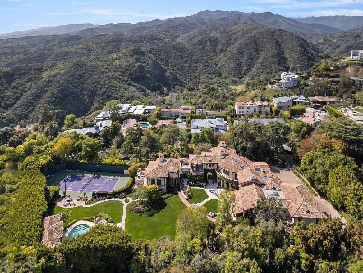 This high-angle aerial view captures a sprawling Mediterranean-style estate nestled within a lush, hilly landscape. The property features a large terracotta-tiled roof, a private tennis court, a swimming pool, and expansive manicured lawns surrounded by dense greenery. The surrounding terrain showcases rolling mountains, providing a secluded and prestigious setting for this luxury residence.