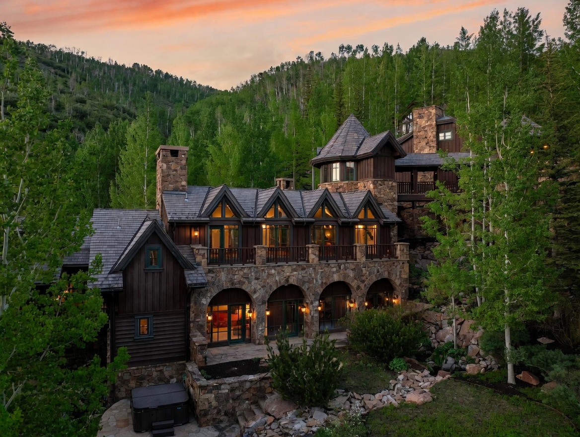 This stunning mountain estate features a multi-level design with a blend of dark wood siding and rugged stone masonry, nestled against a lush, forested hillside. The rear facade showcases a series of arched stone openings on the lower level, a spacious wooden balcony above, and a distinctive turret element, all set against a vibrant sunset sky. A private hot tub is visible on the lower stone patio, emphasizing the home's luxurious and secluded alpine atmosphere.