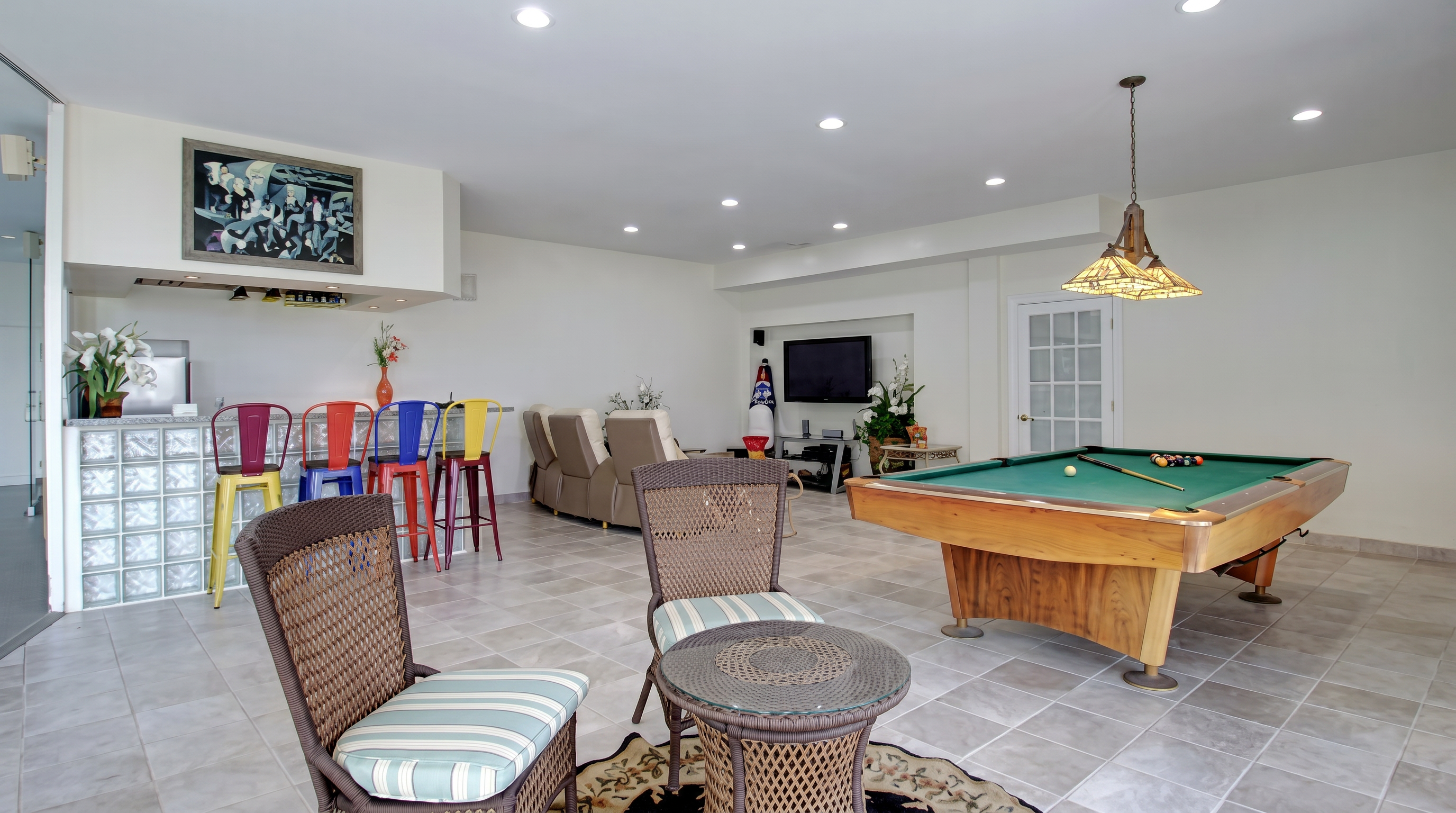 This spacious basement serves as a versatile entertainment area, featuring a pool table, a wet bar with colorful bar stools, and a cozy seating area with a television. The space is finished with light-colored tile flooring and a neutral color palette, creating a relaxed, recreational atmosphere perfect for hosting. Large recessed lights throughout provide bright, even illumination across the open floor plan.