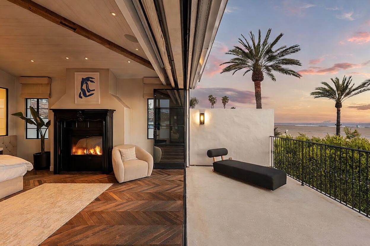 This primary bedroom features a seamless transition to an outdoor balcony through large, retractable glass doors, offering a panoramic view of the ocean at sunset. The interior is anchored by a dark, ornate fireplace surround and rich, herringbone-patterned wood flooring, creating a sophisticated and cozy atmosphere. A minimalist lounge chair sits near the hearth, while a sleek, modern daybed is positioned on the balcony to take full advantage of the coastal scenery.