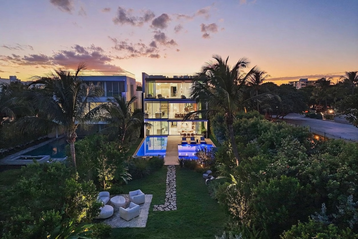 This aerial perspective captures a modern, multi-story residence at dusk, showcasing its sleek, glass-heavy architecture and illuminated interior levels. The property features a private swimming pool with a stone walkway leading through a lush, landscaped lawn, which includes a dedicated outdoor seating area. The warm glow of the sunset provides a dramatic backdrop, highlighting the clean lines and contemporary design of the home.
