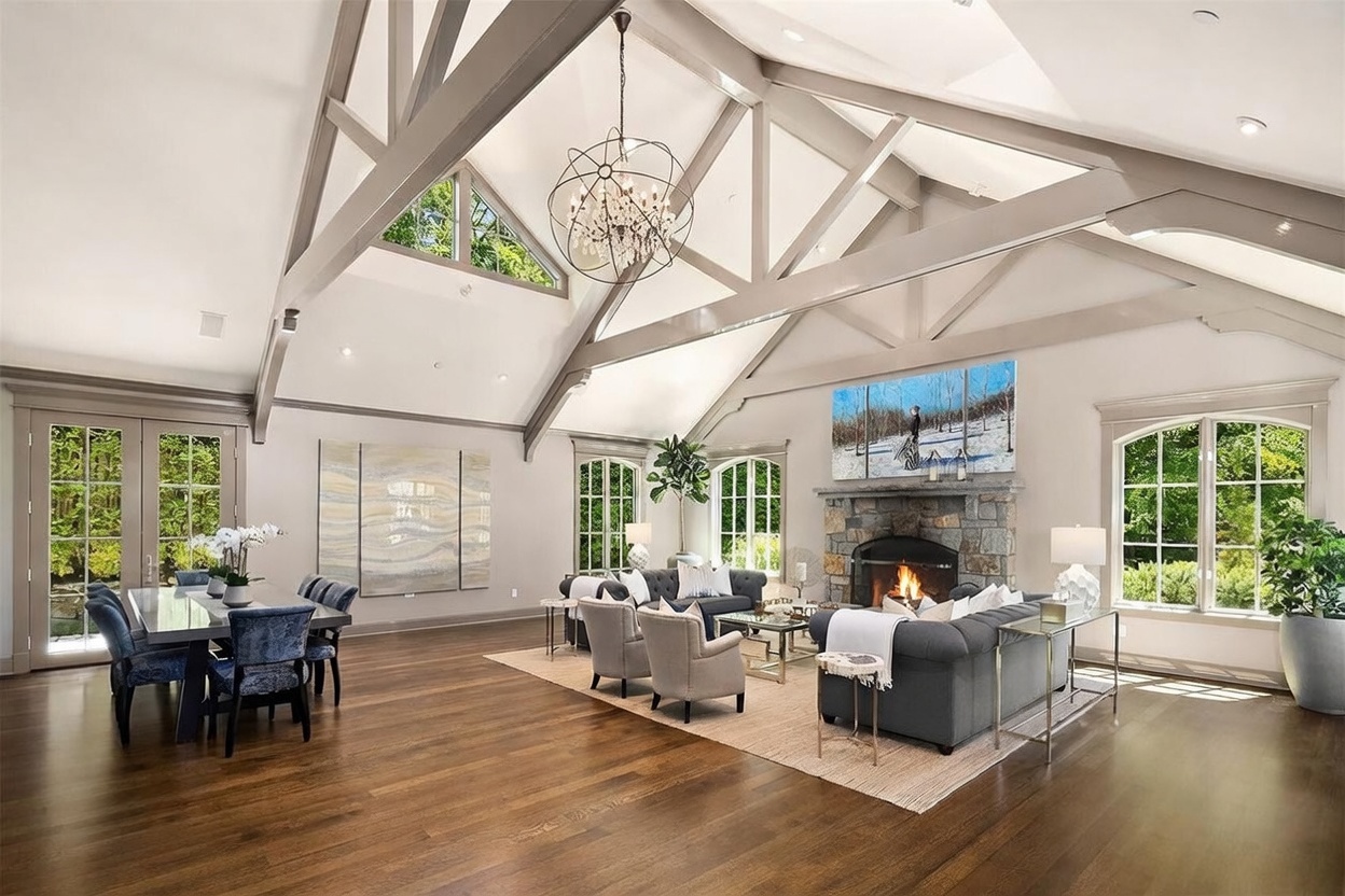 This expansive open-concept living and dining area features dramatic vaulted ceilings with exposed structural beams and a large, intricate chandelier. A stone-clad chimney breast serves as the focal point of the living space, which is furnished with plush grey seating arranged on a textured area rug. The room is bathed in natural light from multiple tall windows and glass doors, creating an airy and sophisticated atmosphere.