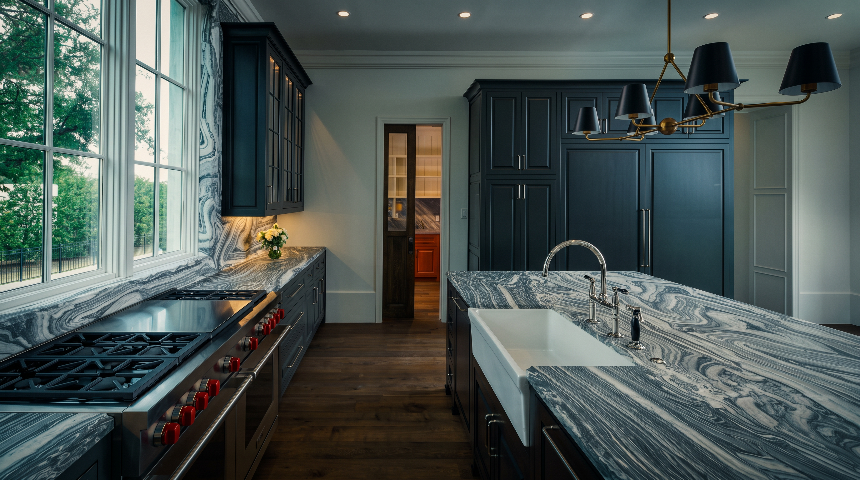 This sophisticated kitchen features dark cabinetry paired with striking, heavily veined stone countertops and backsplashes that create a dramatic visual impact. A large central island houses a farmhouse-style sink, while a professional-grade range with red knobs serves as a focal point along the perimeter. The space is anchored by warm-toned wood flooring and illuminated by a modern, multi-arm chandelier, offering a view through a doorway into an adjacent room.