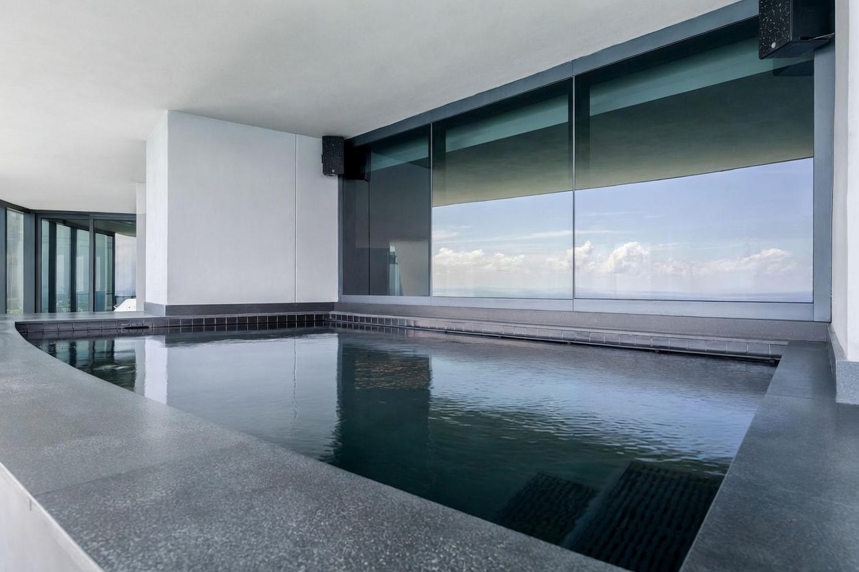 This image captures a sleek, modern plunge pool situated on a high-rise balcony, featuring dark, minimalist tiling and a clean-lined design. The pool is framed by expansive, floor-to-ceiling glass panels that reflect the sky and clouds, creating a seamless transition between the water and the horizon. The perspective is low and intimate, emphasizing the tranquil atmosphere and the sophisticated architectural integration of the water feature within the living space.