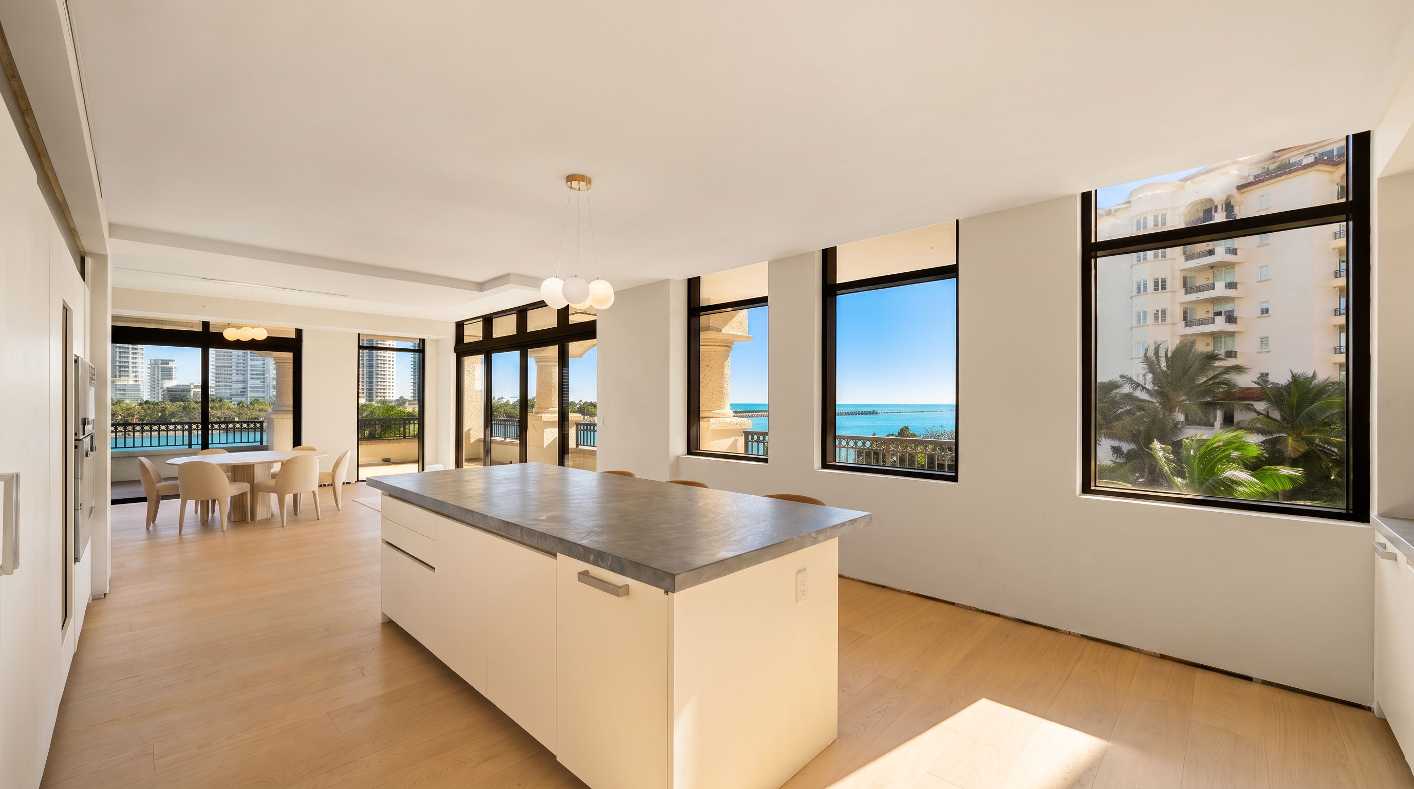 This bright, open-concept kitchen features a large central island with a dark stone countertop and light-toned cabinetry. The space is illuminated by natural light streaming through expansive windows that offer panoramic views of the ocean and surrounding buildings. A dining area is visible in the background, creating a seamless flow between the cooking and eating spaces.