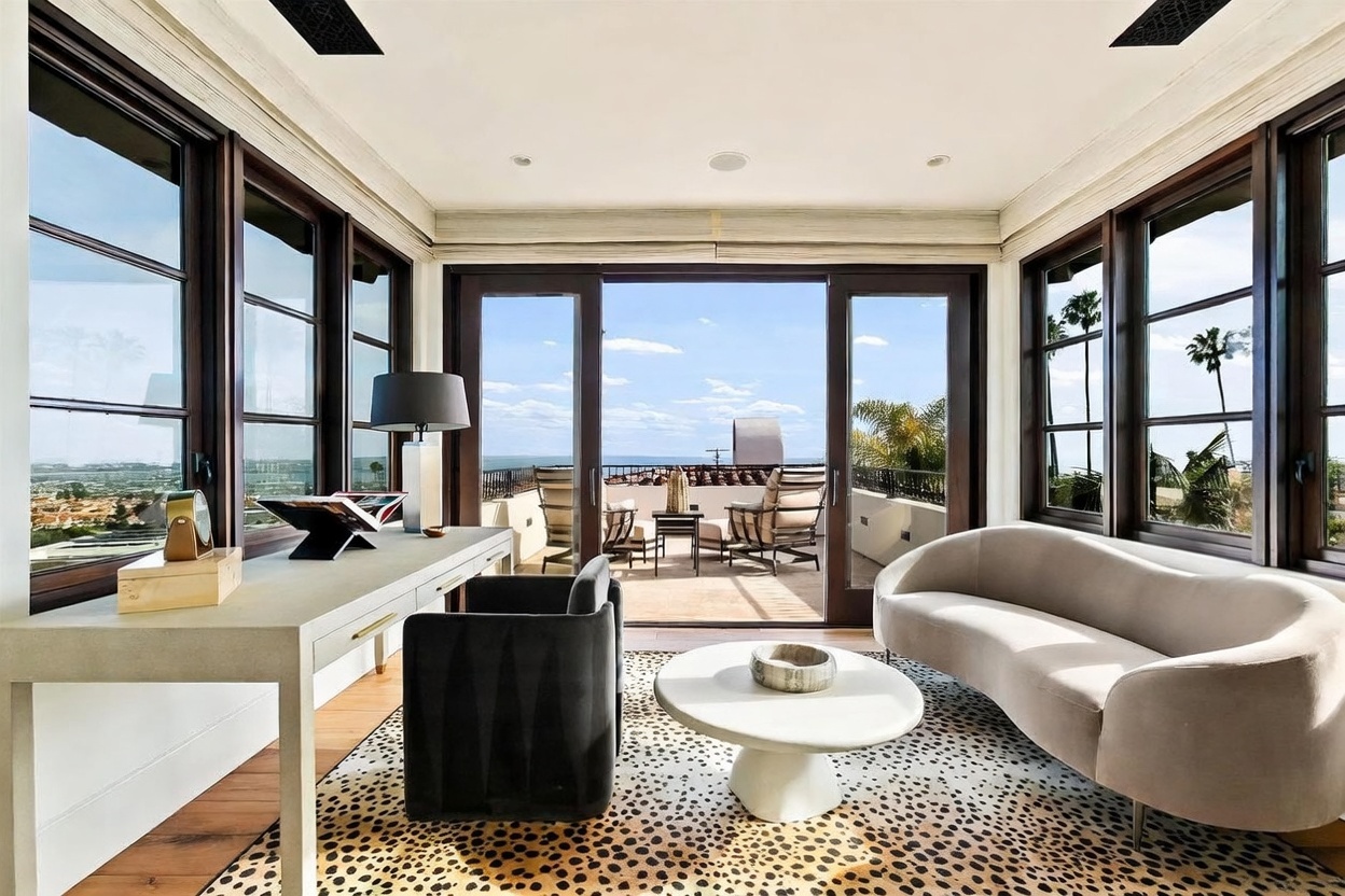 This sophisticated home office features a sleek, light-toned desk paired with a dark upholstered chair, set against a backdrop of large windows that frame a panoramic coastal view. A curved, light-colored sofa and a round coffee table sit atop a bold leopard-print rug, creating a stylish seating area. The room opens directly onto a sun-drenched balcony, seamlessly blending indoor workspace with an inviting outdoor lounge area.