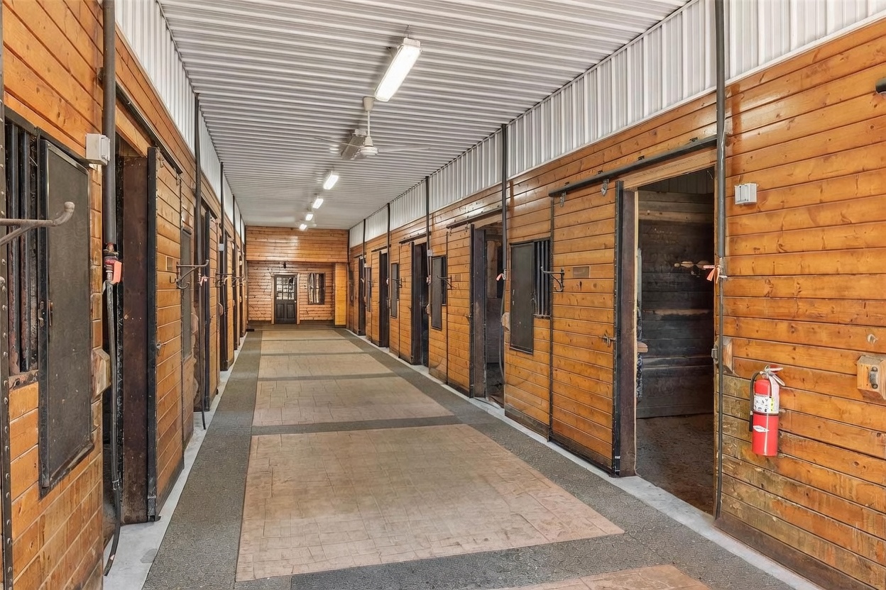 This image shows the interior aisle of a professional horse stable, featuring rows of wooden stalls with sliding doors on either side. The space is illuminated by long, linear overhead lights and a ceiling fan, with a durable, textured floor running the length of the corridor. A fire extinguisher is mounted on the wall near the foreground, and the perspective looks down the center of the aisle toward a door at the far end.