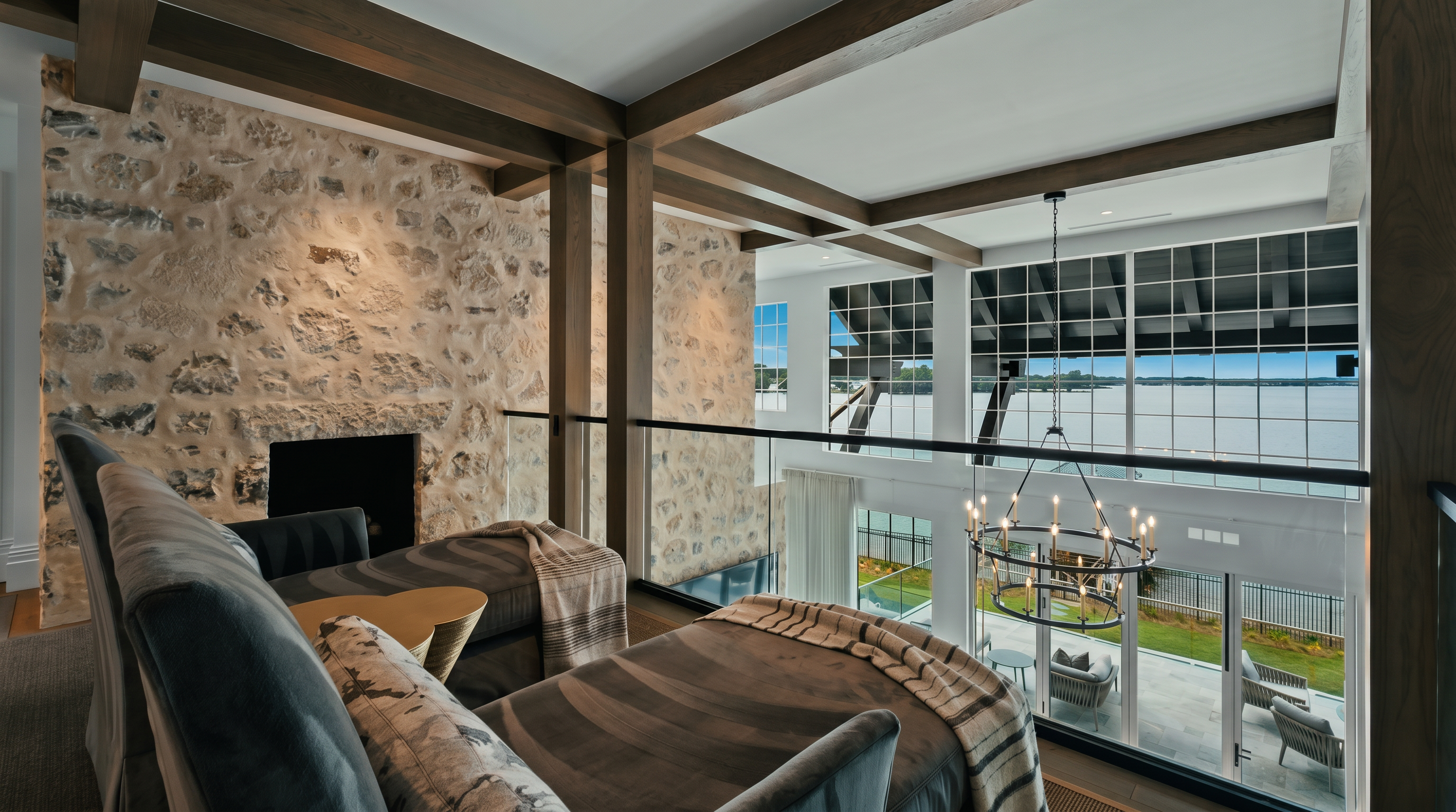 This elevated loft space overlooks a grand living area, featuring a dramatic stone-clad chimney breast that rises through the open-concept design. The room is furnished with two plush, patterned chaise lounges positioned to enjoy the view through expansive windows that look out over a lake. The architectural style is rustic yet refined, characterized by exposed wooden beams and a glass railing that maintains an airy, open connection to the level below.