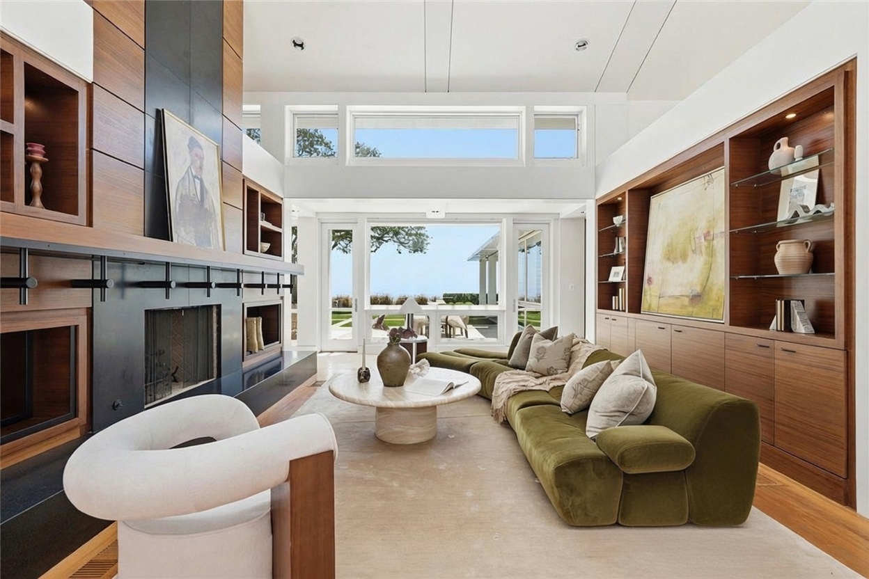 This sophisticated living room features a large, olive-green sectional sofa and a unique, sculptural white armchair arranged around a circular stone coffee table. A prominent, dark-toned fireplace surround serves as a focal point, complemented by extensive built-in wood shelving and cabinetry on the opposite wall. Large windows and glass doors provide an expansive view of the outdoor landscape, bathing the room in natural light and creating a seamless connection to the exterior.