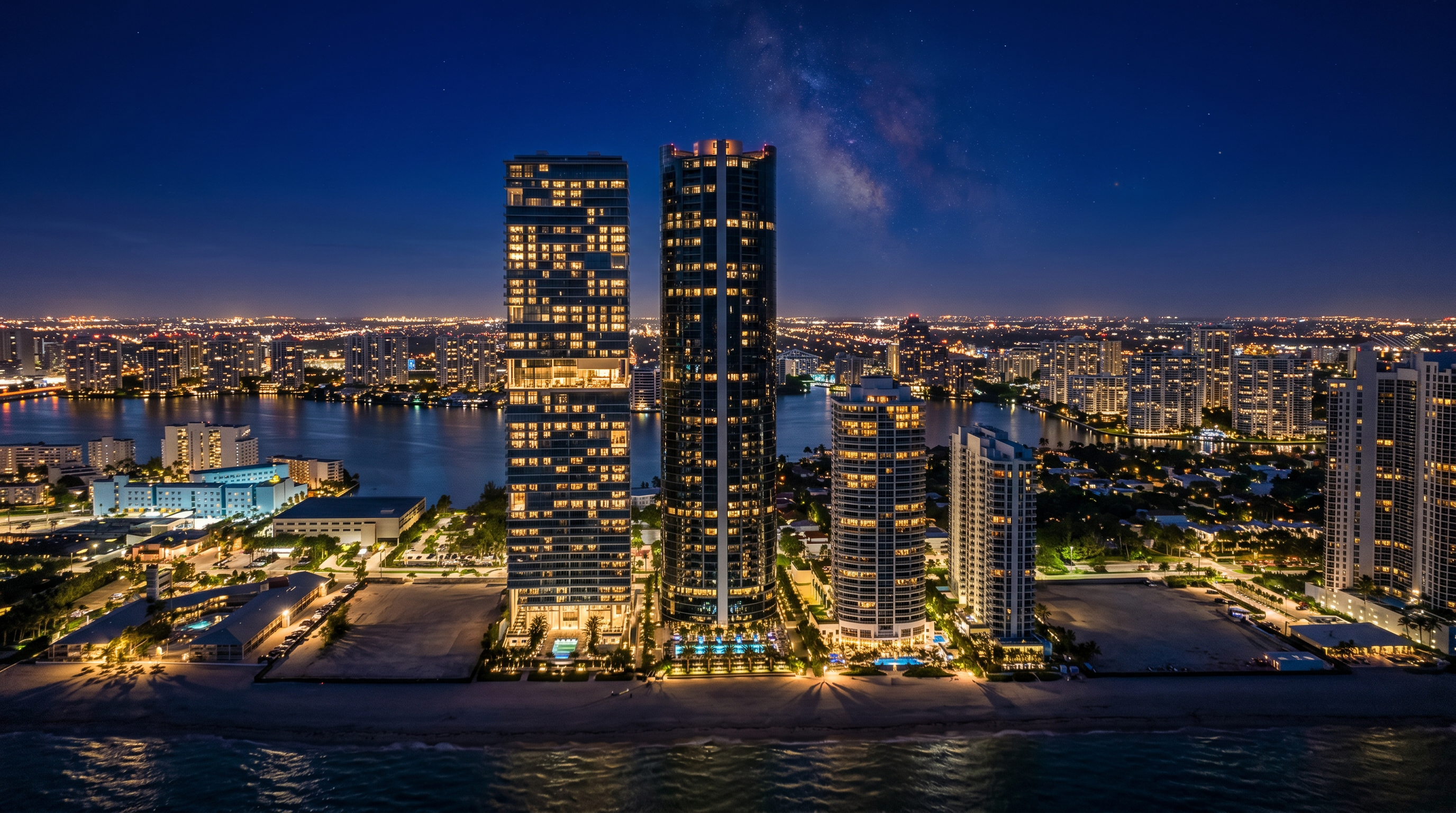 This high-altitude aerial view captures a prominent stretch of coastline featuring several modern high-rise residential towers situated directly along a pristine sandy beach. The composition highlights the contrast between the deep blue ocean, the expansive beach, and the dense urban development of the surrounding city skyline. The perspective provides a comprehensive look at the property's prime waterfront location and its integration into the coastal landscape.