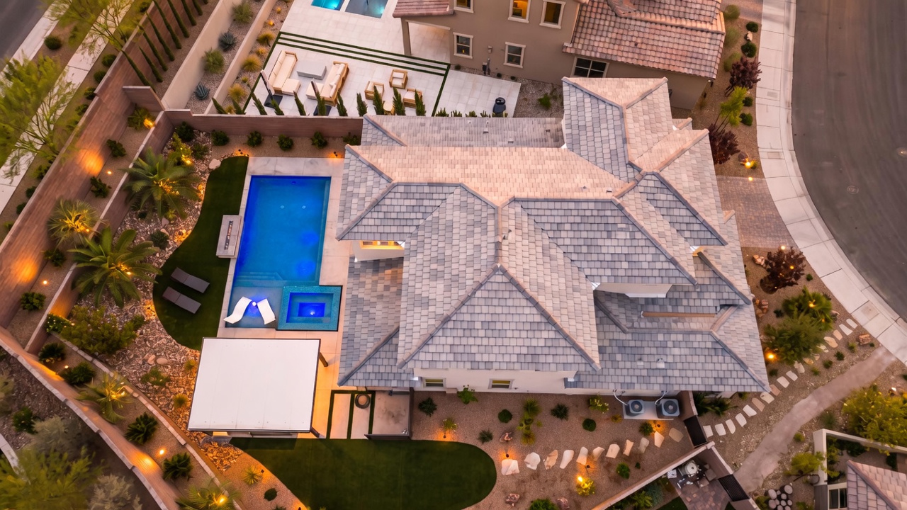 This high-angle aerial view captures a modern residential property featuring a complex, multi-faceted gray tiled roof. The backyard is meticulously landscaped with a vibrant blue swimming pool, a built-in spa, a covered patio area, and a mix of artificial turf and desert-style gravel beds. The perspective provides a comprehensive look at the home's footprint and the surrounding outdoor living spaces, highlighting the clean, contemporary design of the property.