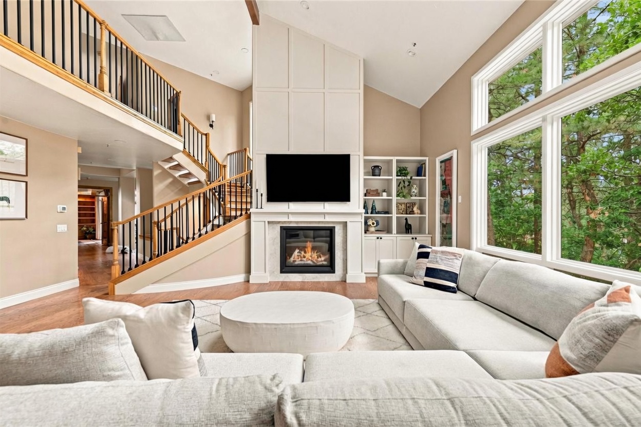 This spacious living room features a vaulted ceiling and a prominent fireplace with a tall, paneled surround that serves as a focal point. A large, light-colored sectional sofa and a round ottoman are arranged on a patterned rug, creating a comfortable seating area, while a staircase with a wooden railing leads to an upper landing. Large windows on the right side of the room provide ample natural light and views of the surrounding trees.