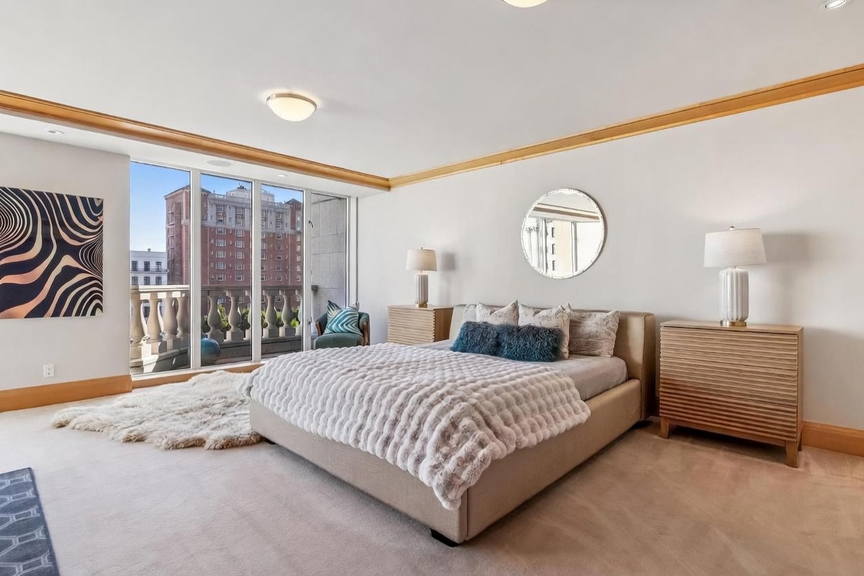 This spacious primary bedroom features a neutral color palette, a plush upholstered bed, and two light wood nightstands topped with matching table lamps. A large circular mirror hangs above the bed, while a textured area rug adds warmth to the carpeted floor. Large windows provide ample natural light and offer a view of the adjacent balcony and city architecture.